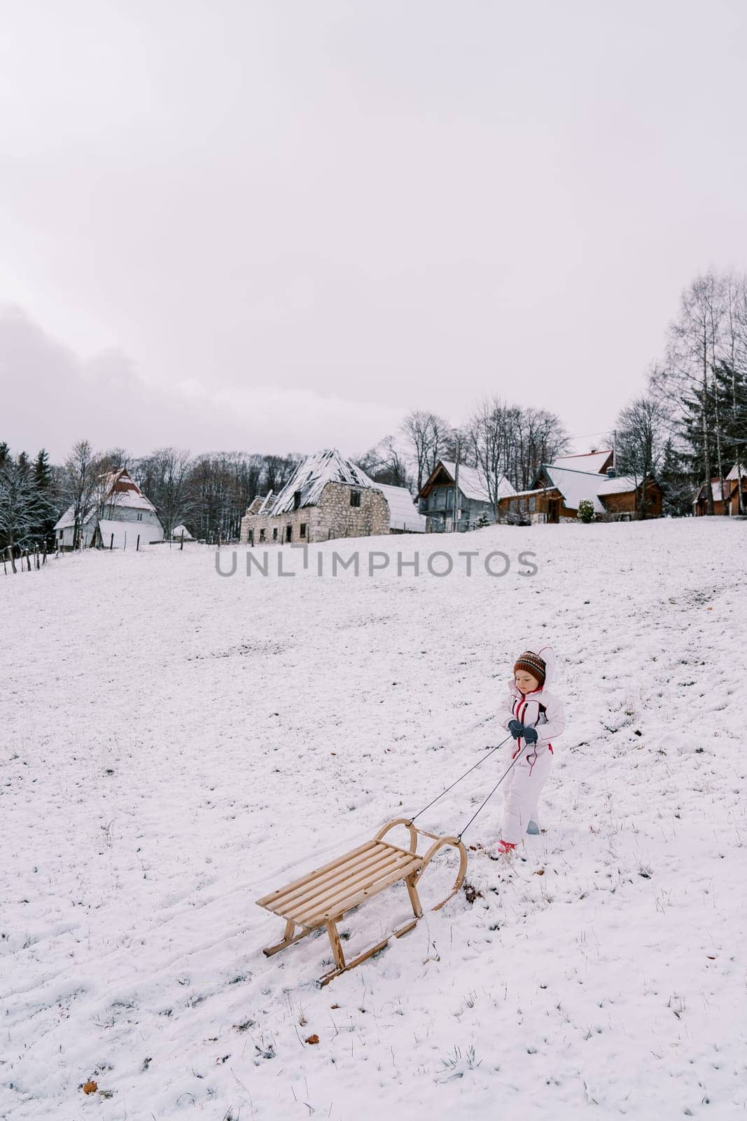 Little girl drags a sled on a string up a hill, looking at it. High quality photo