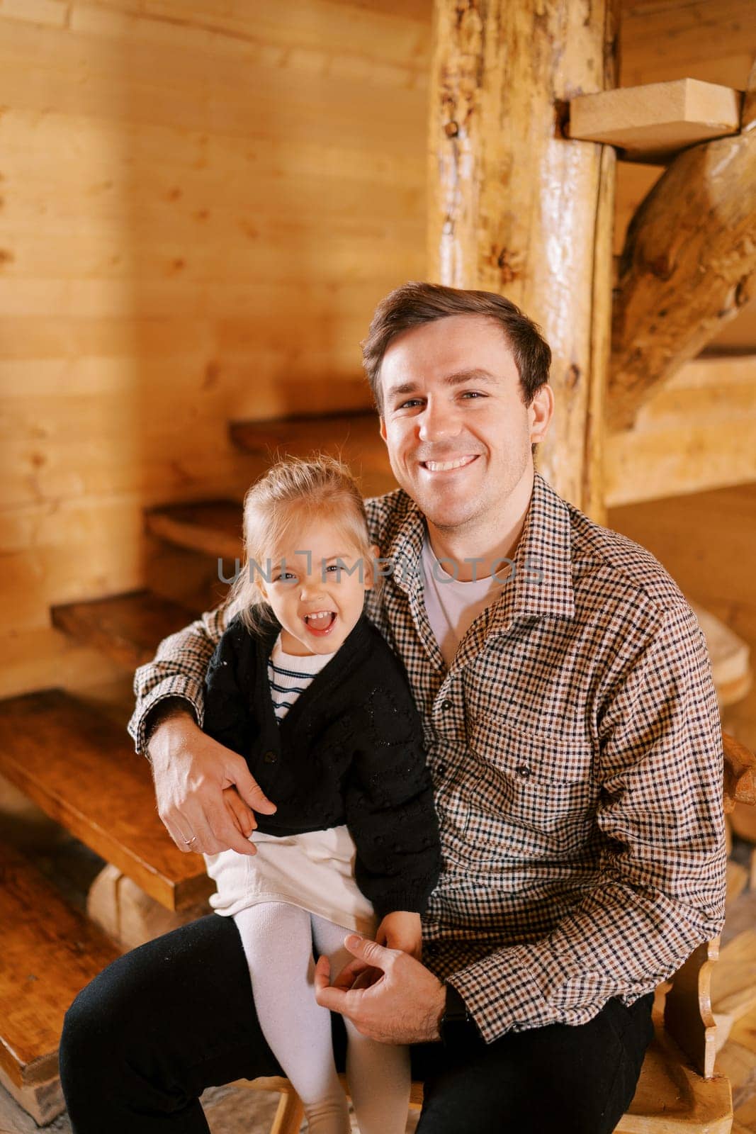 Little laughing girl sitting on smiling dad lap near wooden stairs in cottage. High quality photo