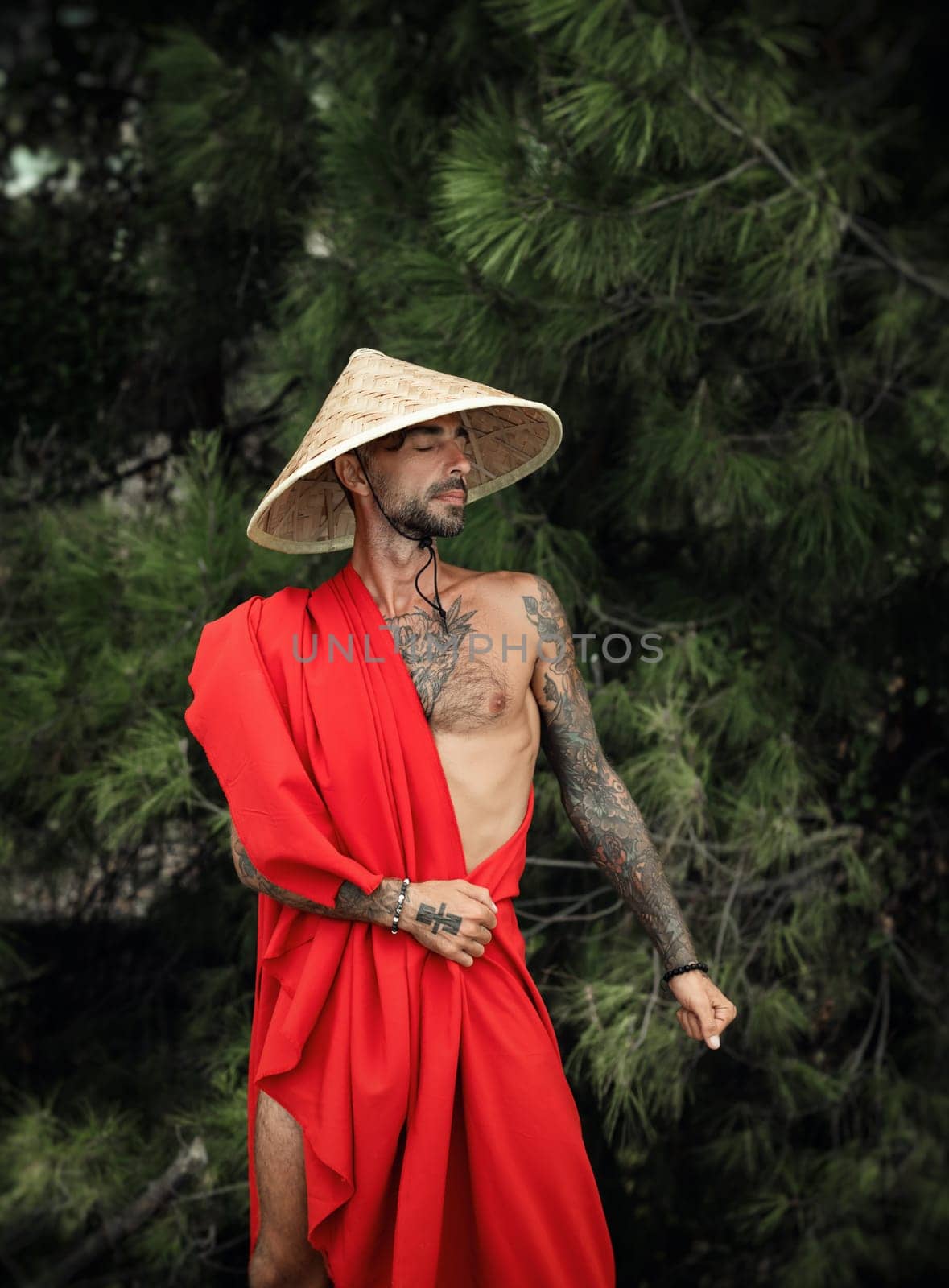 Sexy male monk in red cape and an Asian triangular hat with tattoos in nature by Rotozey