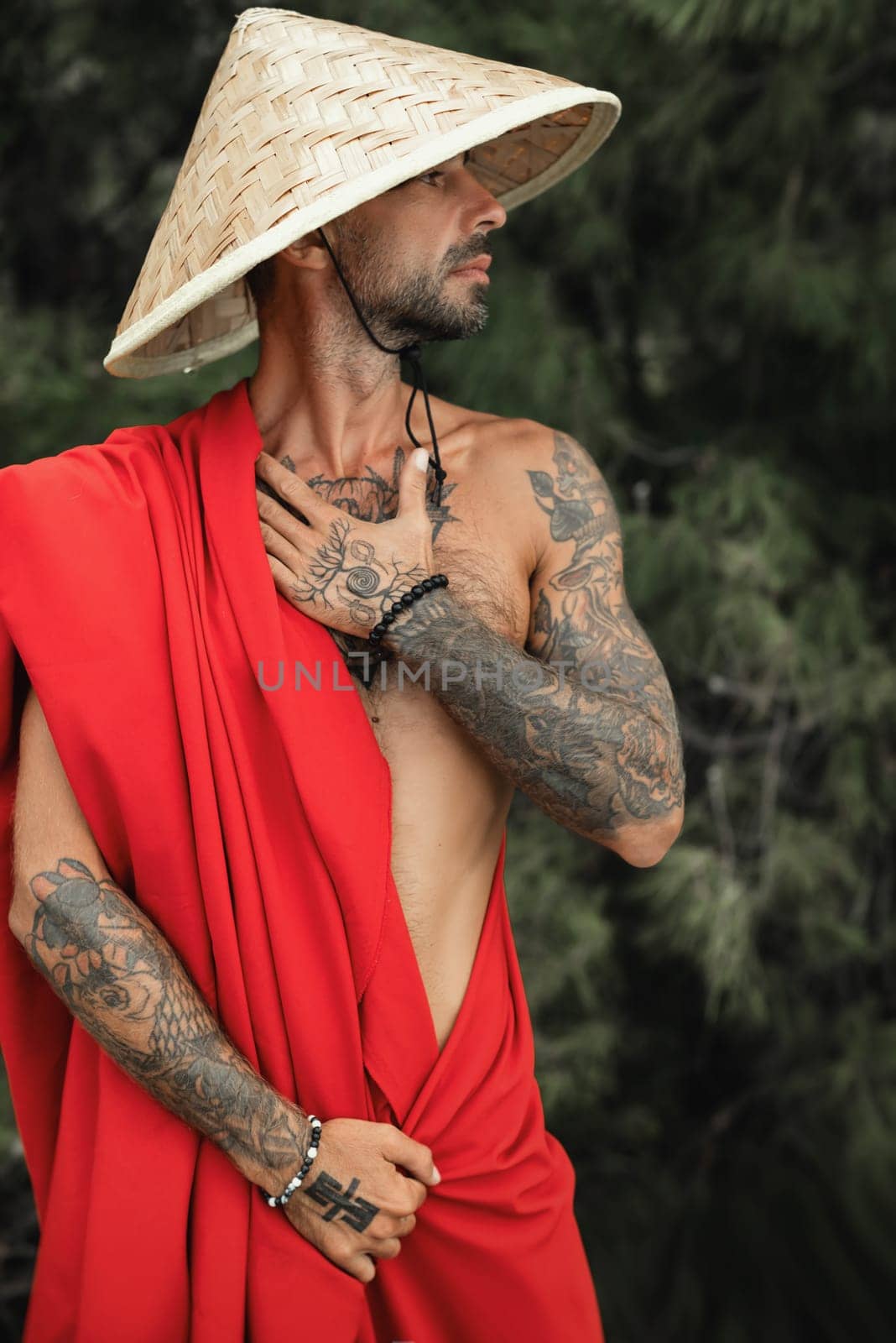 Sexy male monk in a red cape and an Asian triangular hat with tattoos in nature