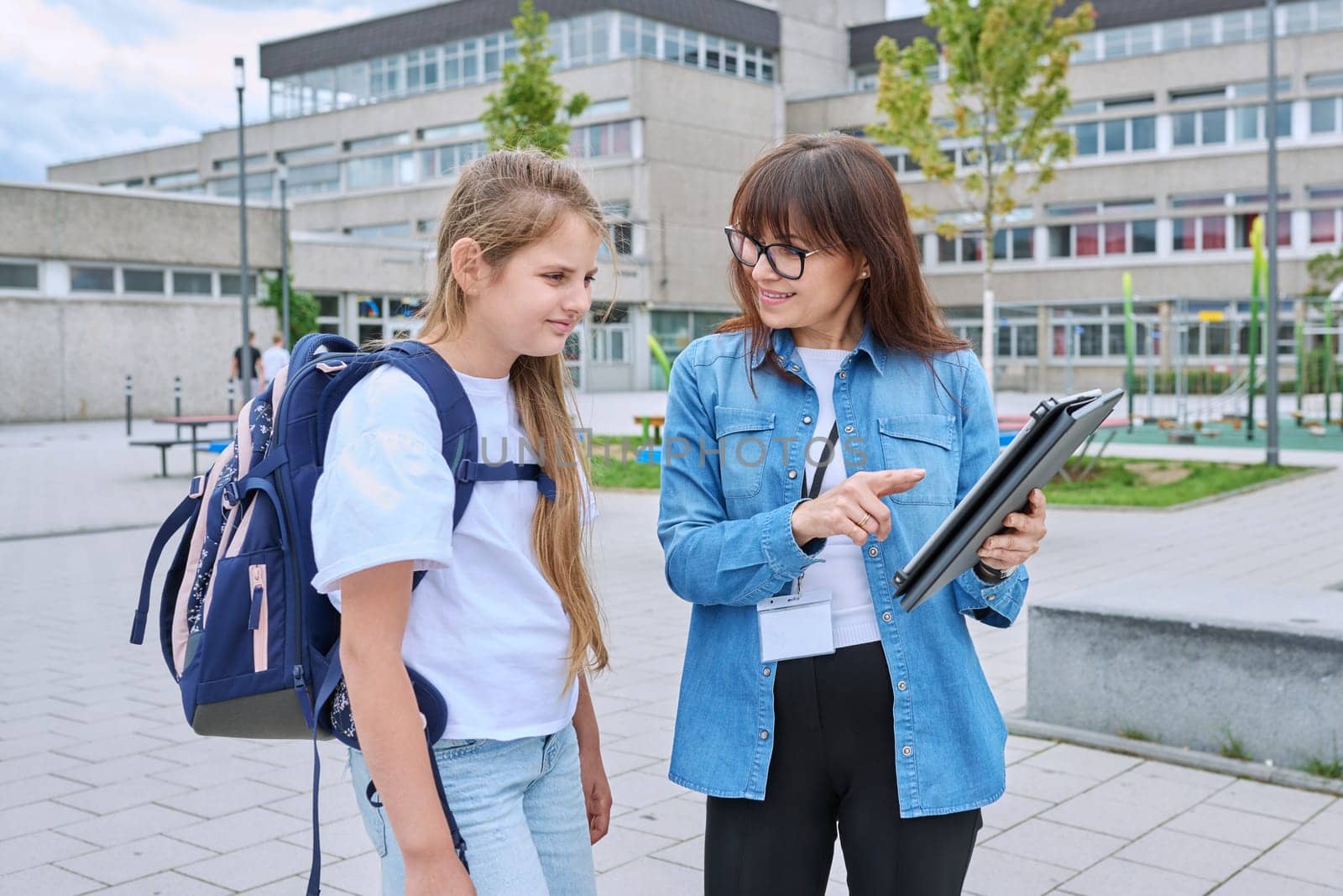 Talking female teacher and schoolgirl child outdoor, school building background. Meeting communication student girl with backpack and mentor counselor. Education, pre-teenage, learning, back to school