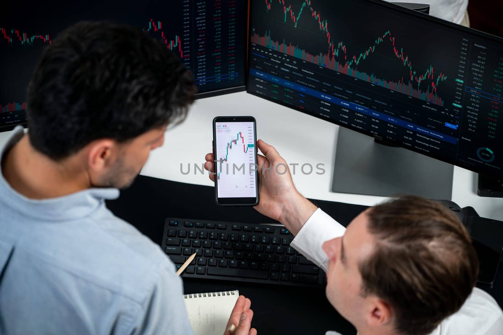 Investor stock officer focusing on dynamic exchange rate database on smartphone, comparing with market dynamic graph with monitor screen. Concept of analysis trading technology investment. Sellable.