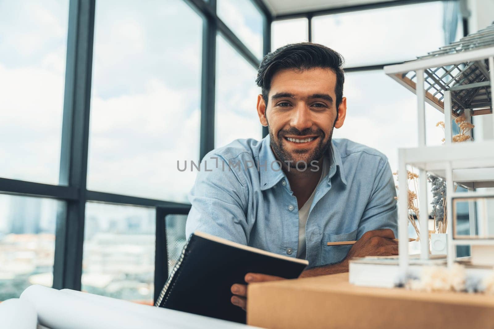 Smiling smart architect engineer looking at camera while sitting near house model. Happy confident businessman wearing casual outfit holding notebook while poses at camera. Civil engineering. Tracery.