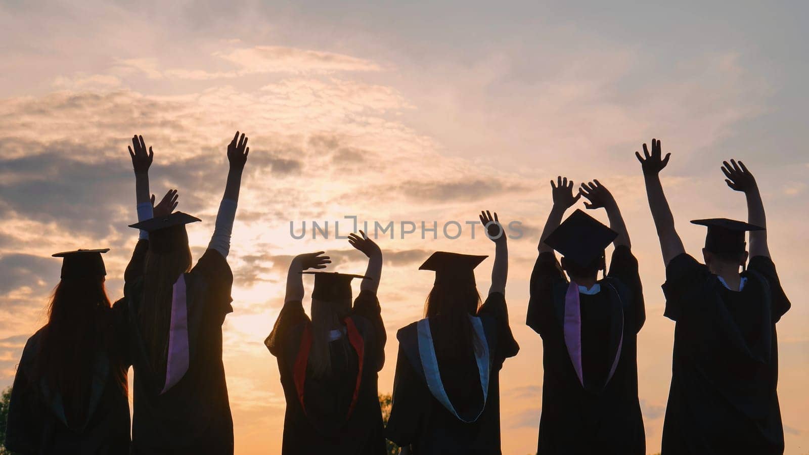 Silhouettes of graduates in black robes waving their arms against the evening sunset. by DovidPro