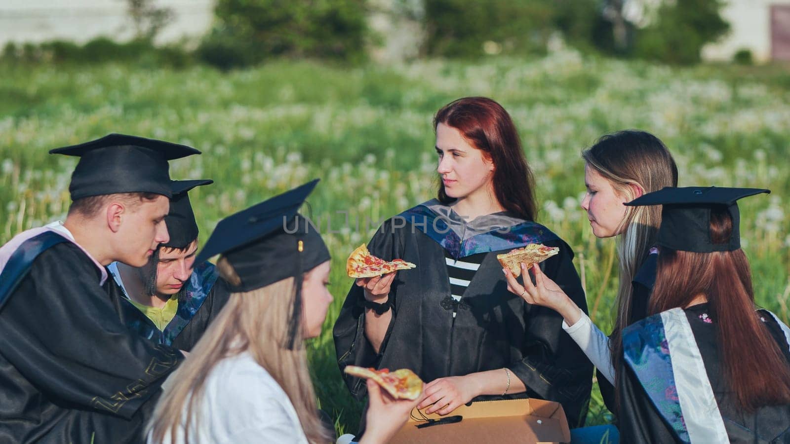 Graduates in black suits eating pizza in a city meadow. by DovidPro