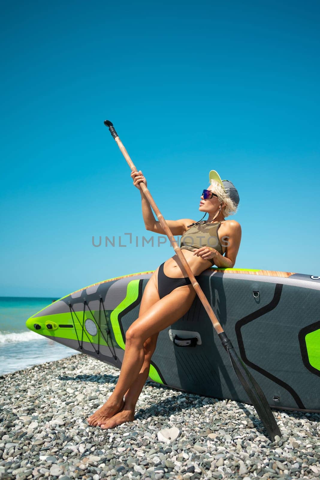 sexy young surfer girl in a swimsuit on a sup board with a bamboo paddle by the sea in stylish sunglasses and a baseball cap