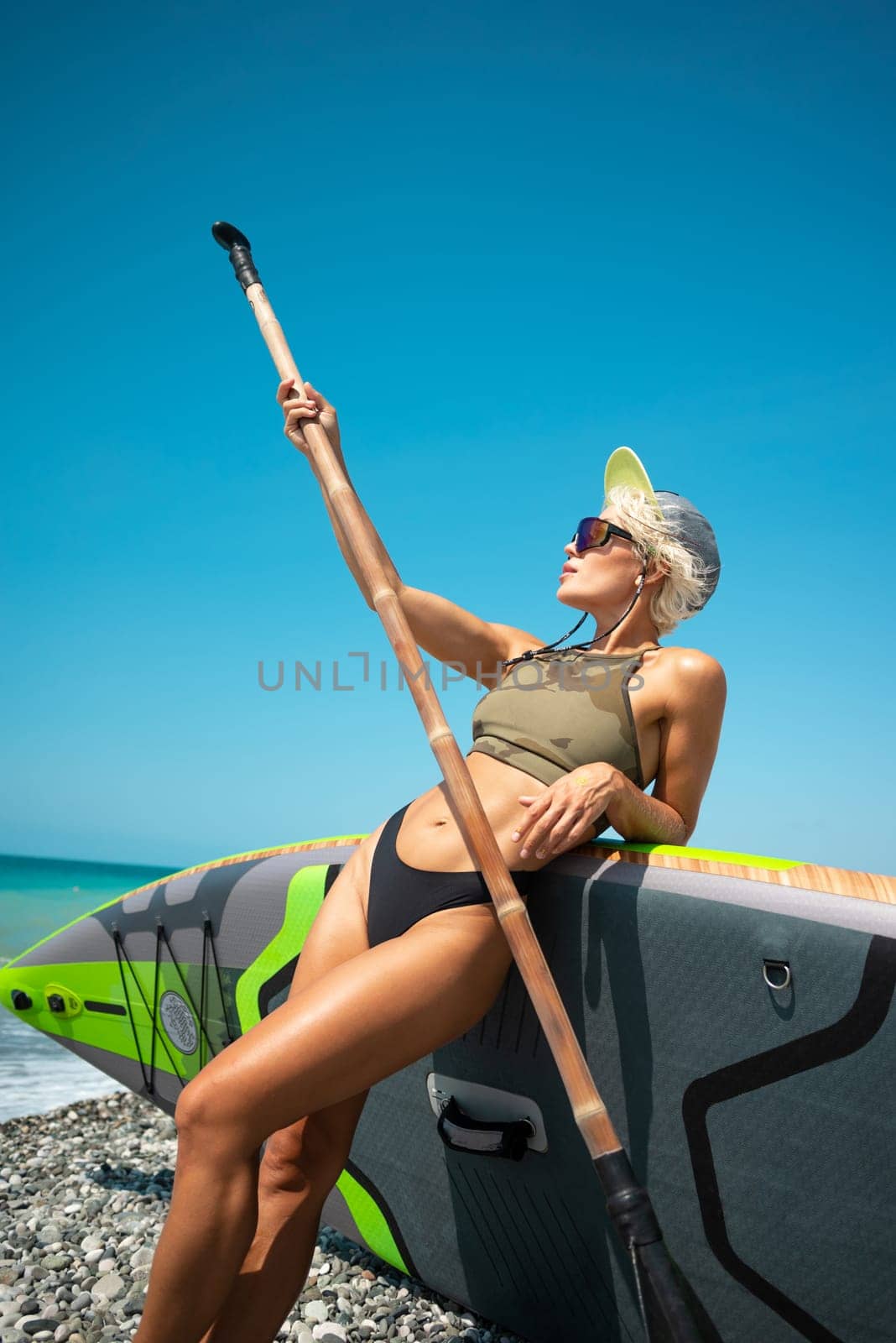 sexy young surfer girl in a swimsuit on a sup board with a bamboo paddle by the sea in stylish sunglasses and a baseball cap