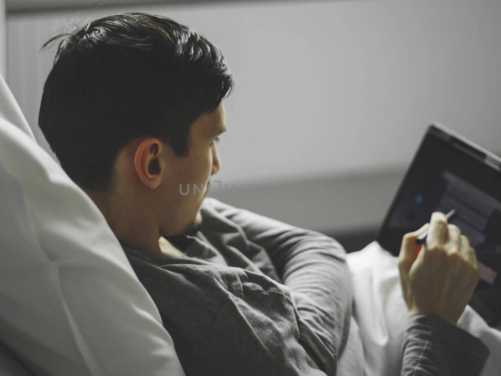 Portrait of one young caucasian guy lying in bed and browsing the news feed on the tablet in the hospital room, close-up side view with selective focus.