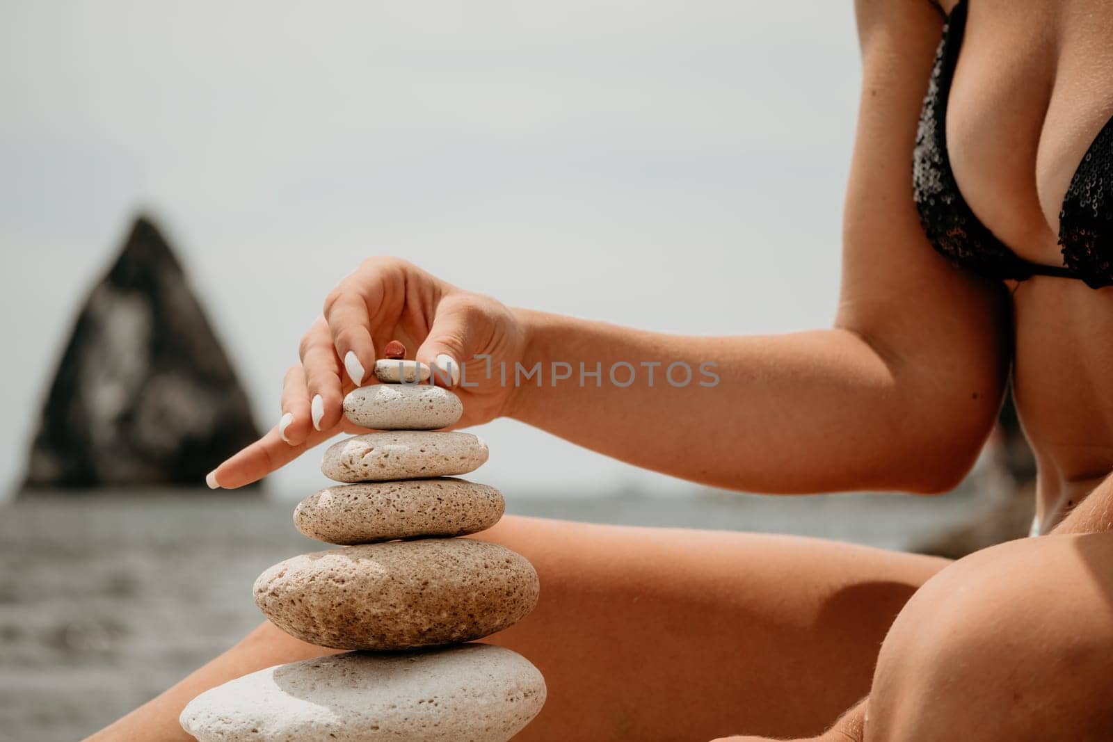 Woman bilds stones pyramid on the seashore on a sunny day on the blue sea background. Happy holidays. Pebble beach, calm sea, travel destination. Concept of happy vacation on the sea, meditation, spa