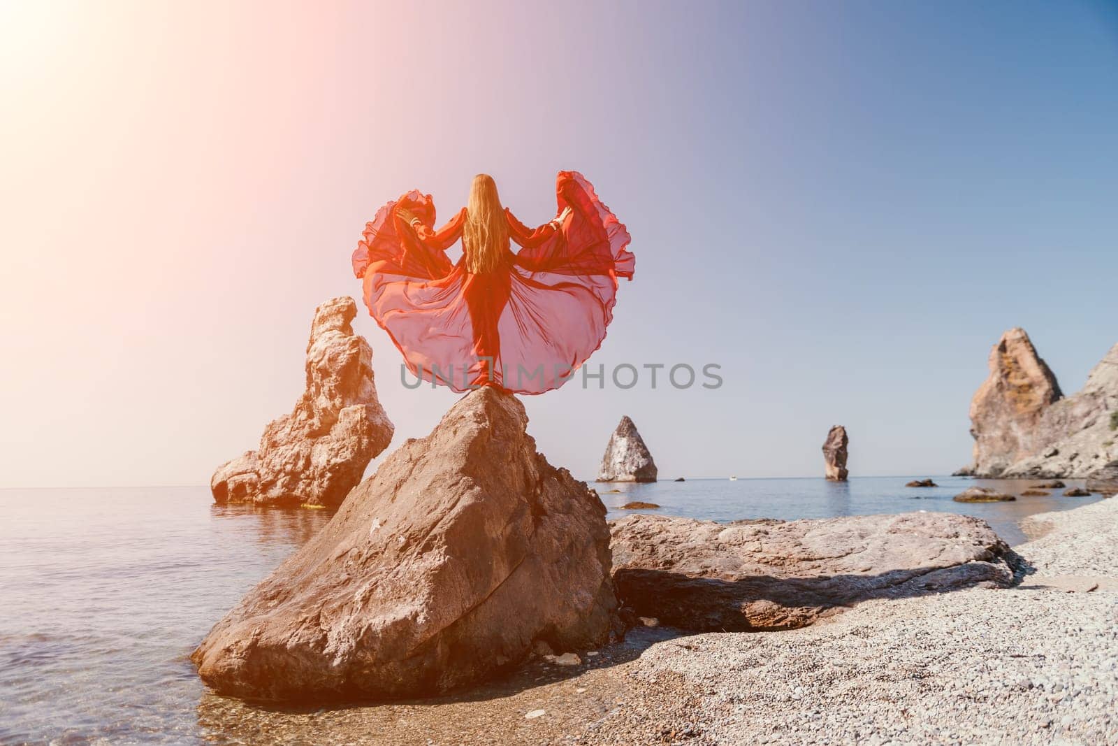 Woman travel sea. Young Happy woman in a long red dress posing on a beach near the sea on background of volcanic rocks, like in Iceland, sharing travel adventure journey by panophotograph