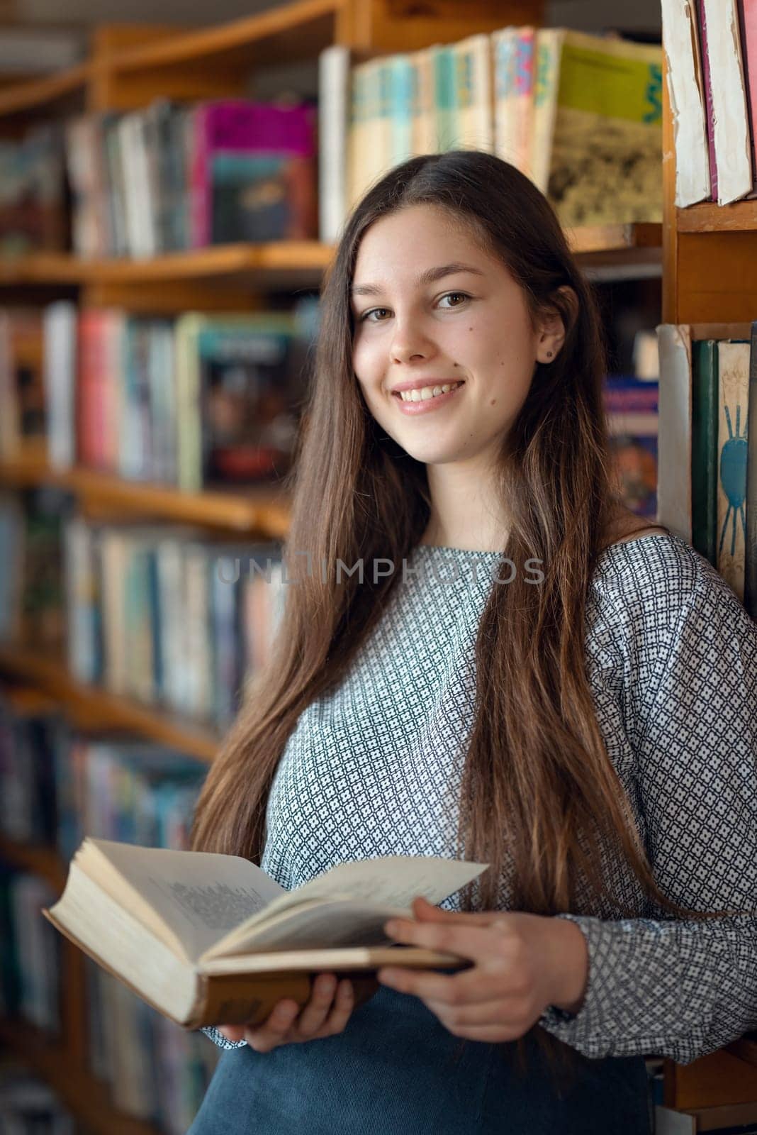 Student with an open book standing near shelves in library, choosing literature for reading