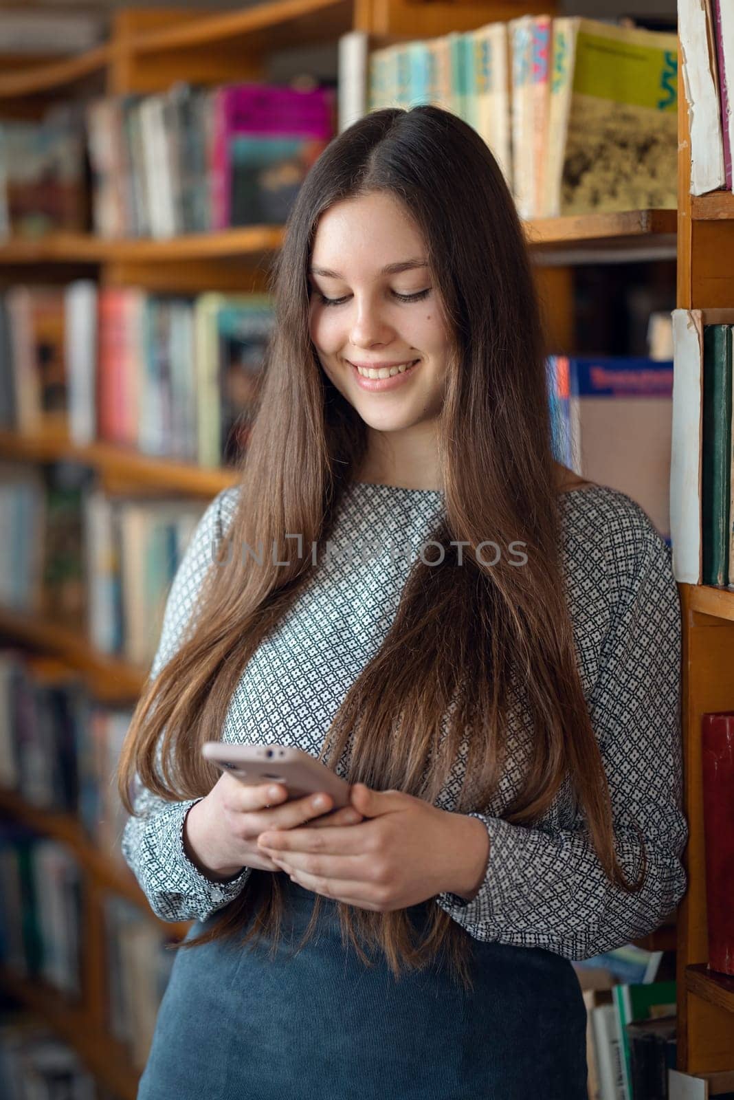 Smiling young girl using phone while being in library, communicating with friends. Students life, girl studying in university library, using mobile phone by VitaliiPetrushenko