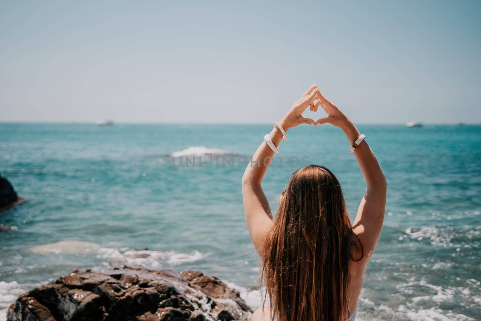 Woman makes heart with hands on beach. Young woman with long hair in white swimsuit and boho style braclets practicing by sea ocean. Concept of longing daydreaming love peace contemplation vacations by panophotograph
