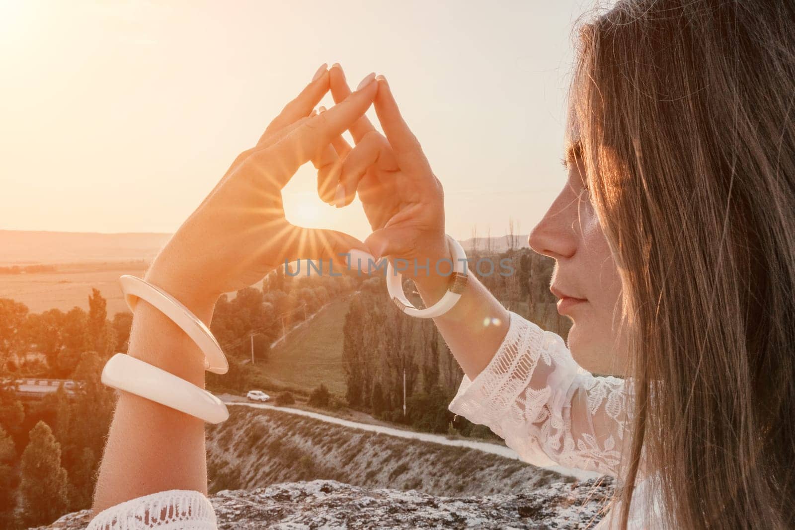 Happy woman in white boho dress making heart sign with hands on sunset in mountains. Romantic woman with long hair standing with her back on the sunset in nature in summer with open hands. by panophotograph