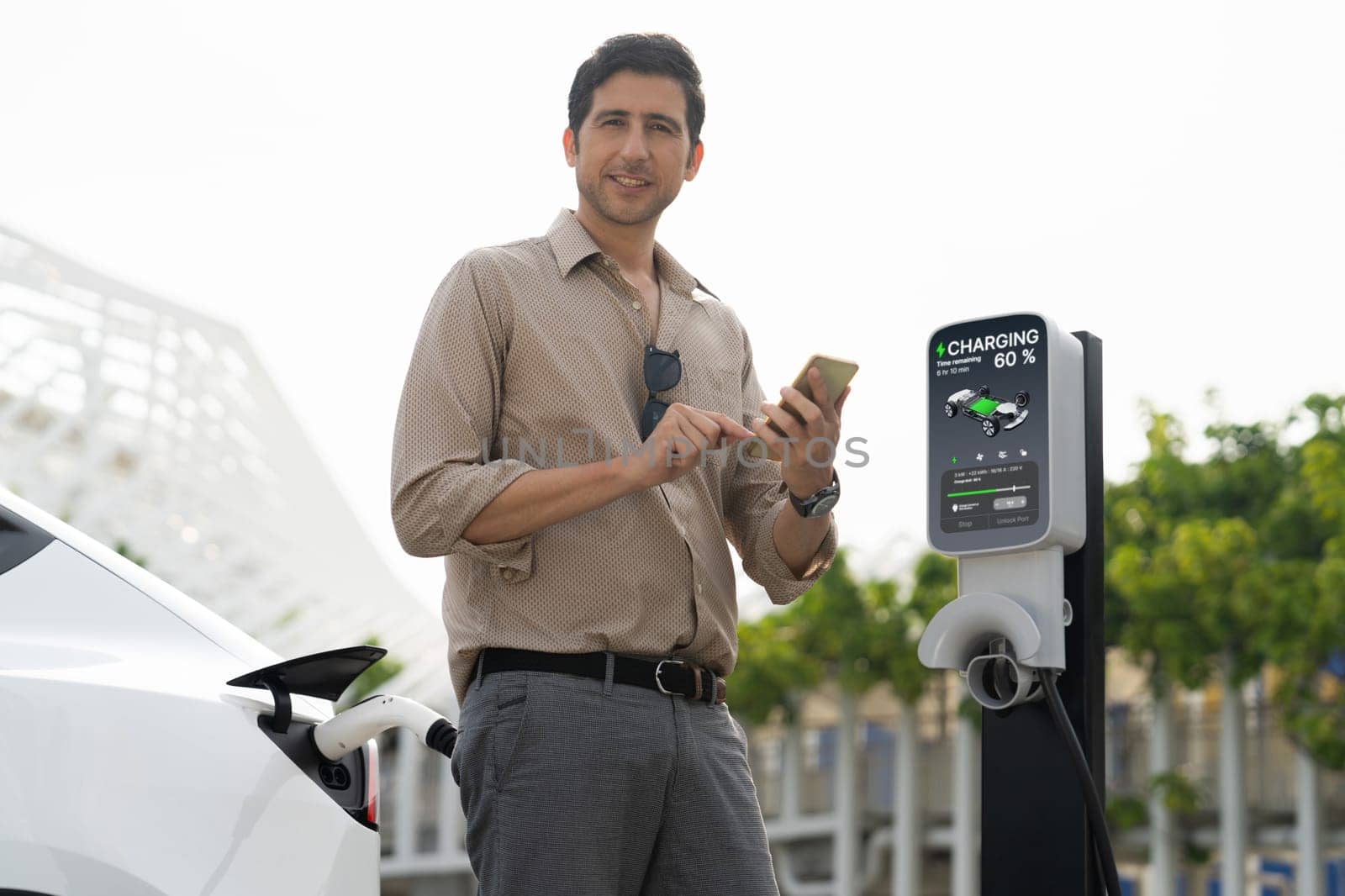 Young man use smartphone to pay for electricity at public EV car charging station green city park. Modern environmental and sustainable urban lifestyle with EV vehicle. Expedient