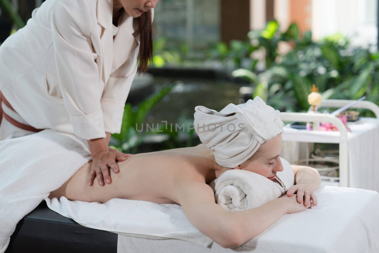 Beautiful young woman received a back massage on a spa bed from masseuse. Attractive female relaxes deeply by skilled hands of the massage therapist. Surrounded with nature. Side view. Tranquility.