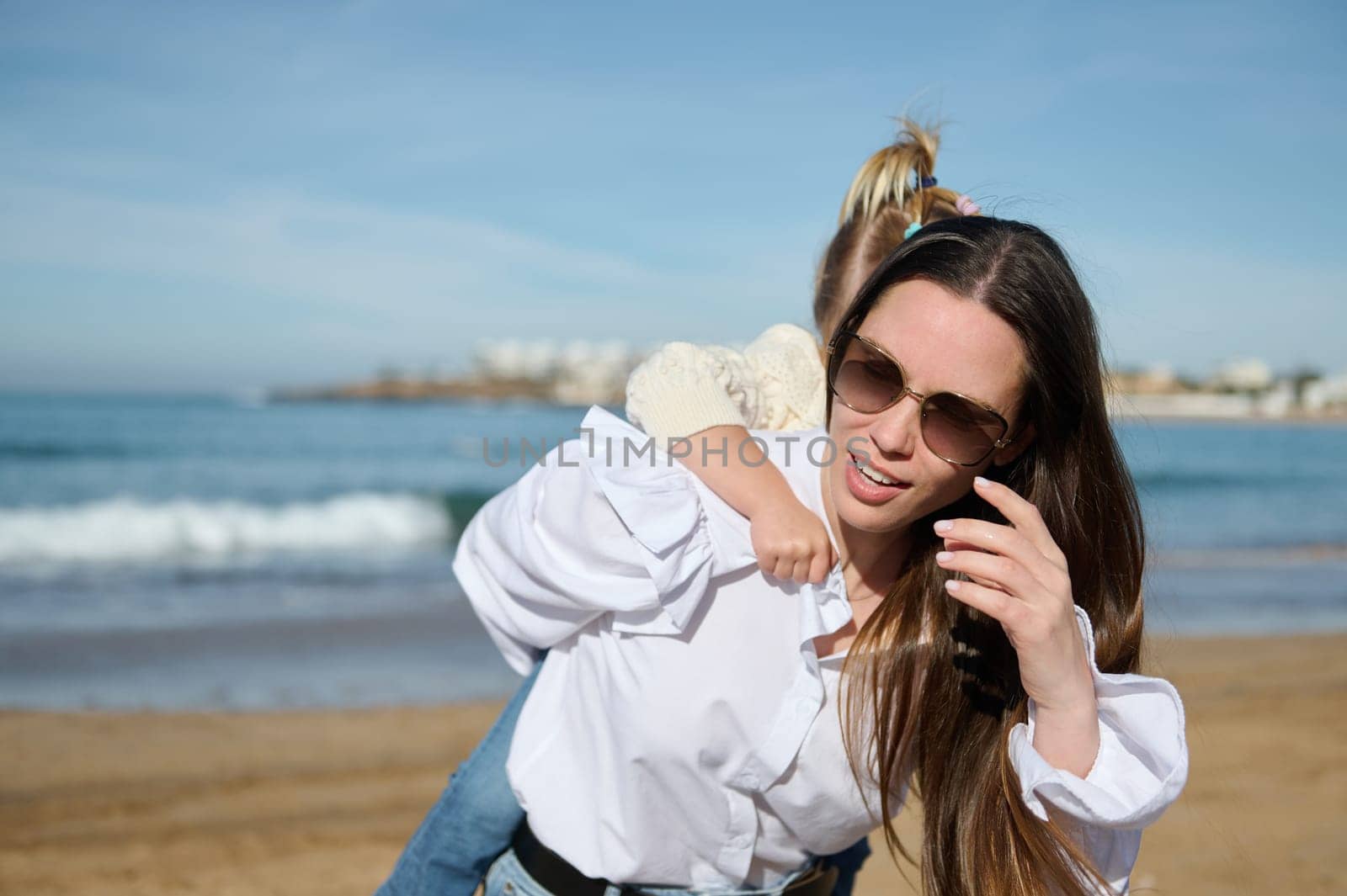 Beautiful woman, young loving caring mother in eyeglasses, carrying her little daughter on her back while spending happy time together on the beach. Family. Motherhood. Carefree childhood