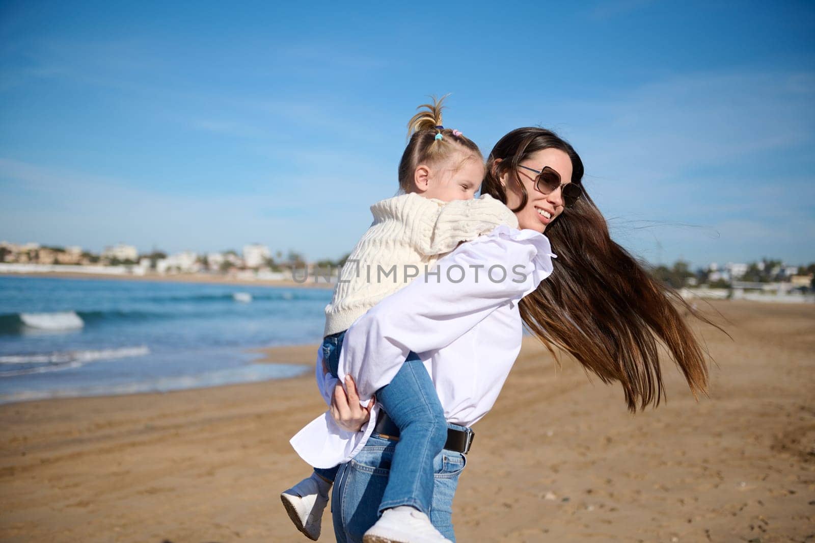Rear view of a happy mother giving piggyback ride to her lovely daughter at beach. Caring mom spinning her child she's holding on her back, enjoying wonderful moments together. Family relationships