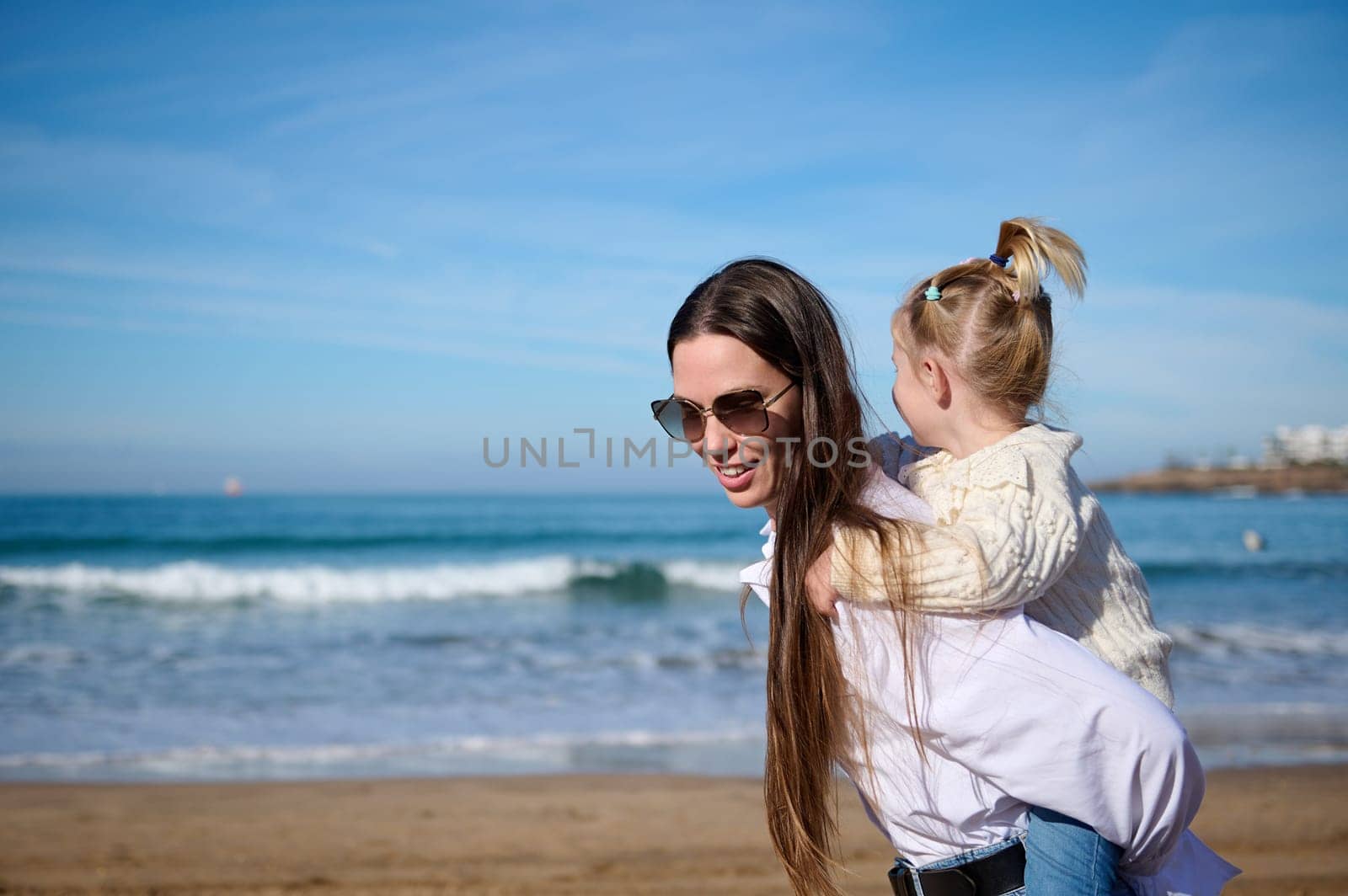 Mom and child girl kid enjoy and fun outdoor lifestyle on the beach. Mother giving piggyback to her lovely daughter by artgf