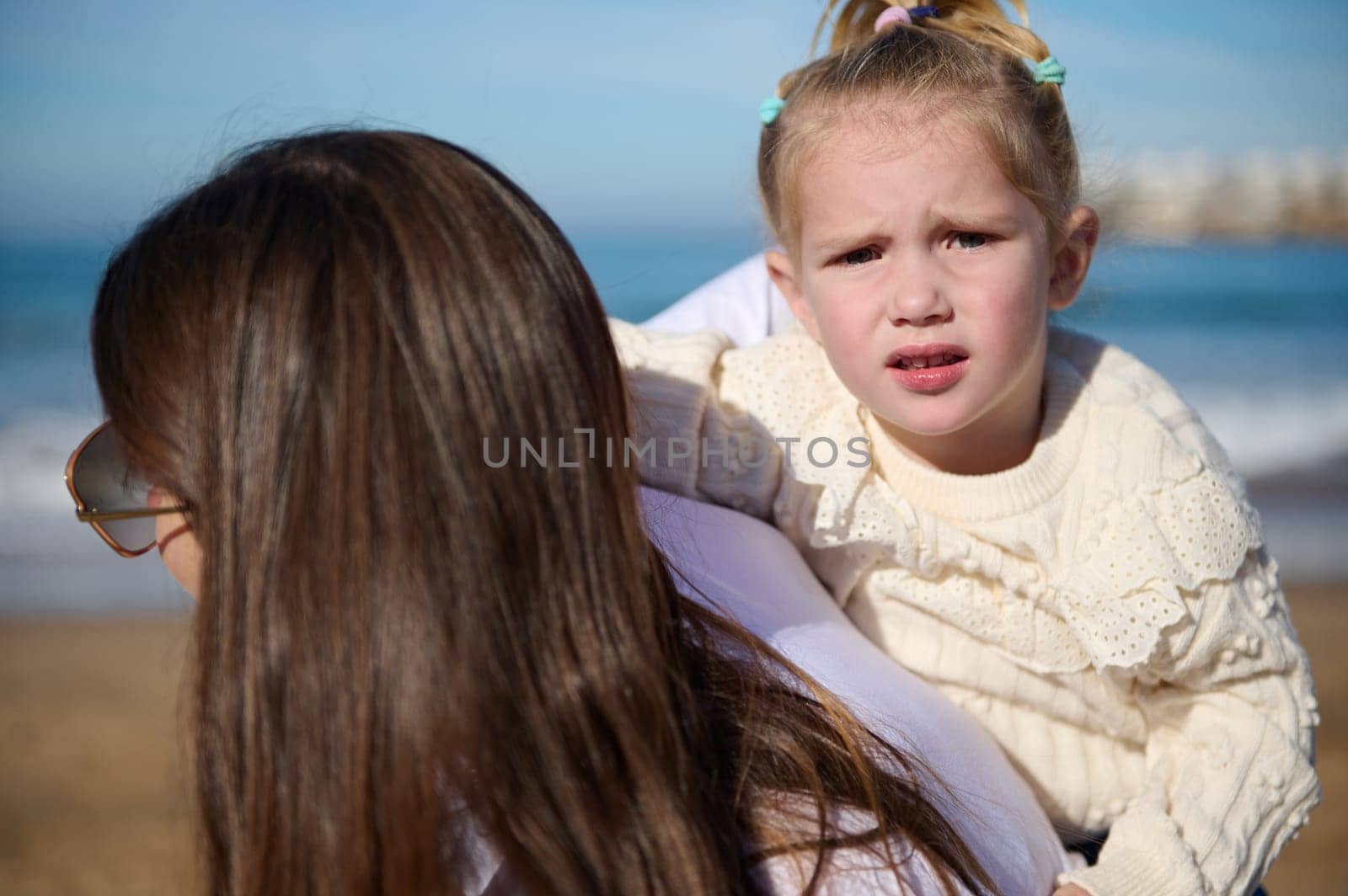 Beautiful Caucasian little child girl looking at the camera while her loving mother carrying her on her back, enjoying walking together on the beach on warm sunny day. People and leisure