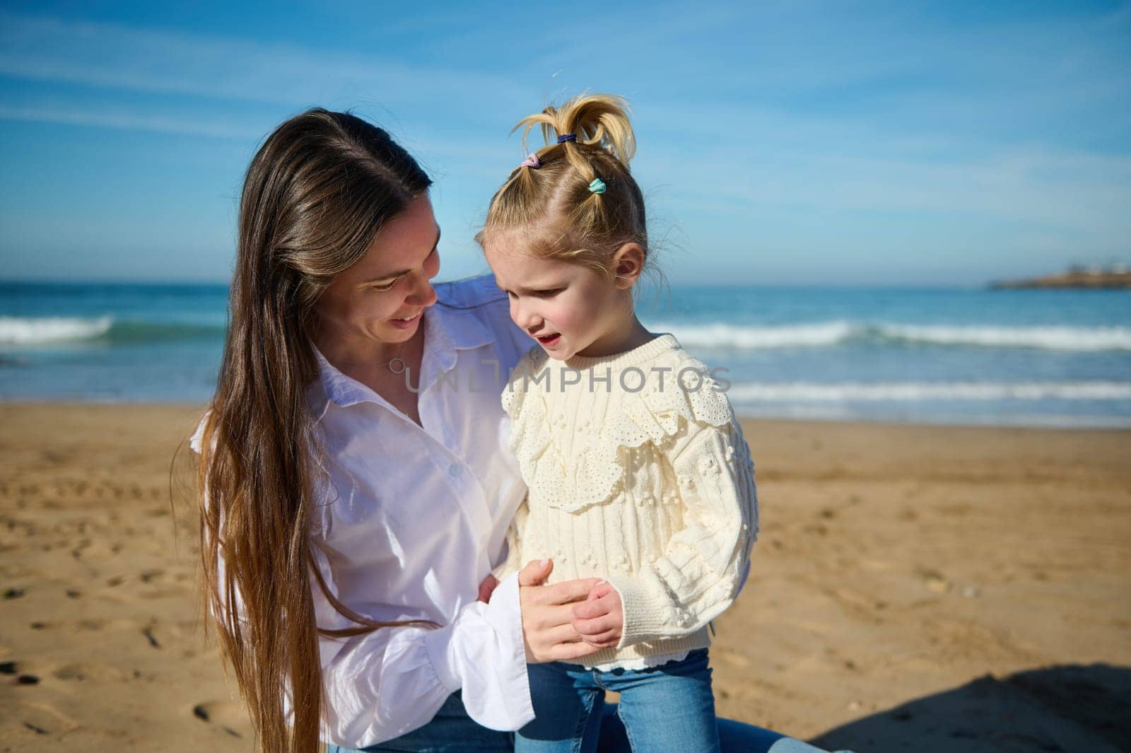 Mother and daughter spending happy time together on the beach on beautiful warm sunny day, sitting on the sandy beach against waves splashing while crashing on the Atlantic shore. Family. Childhood