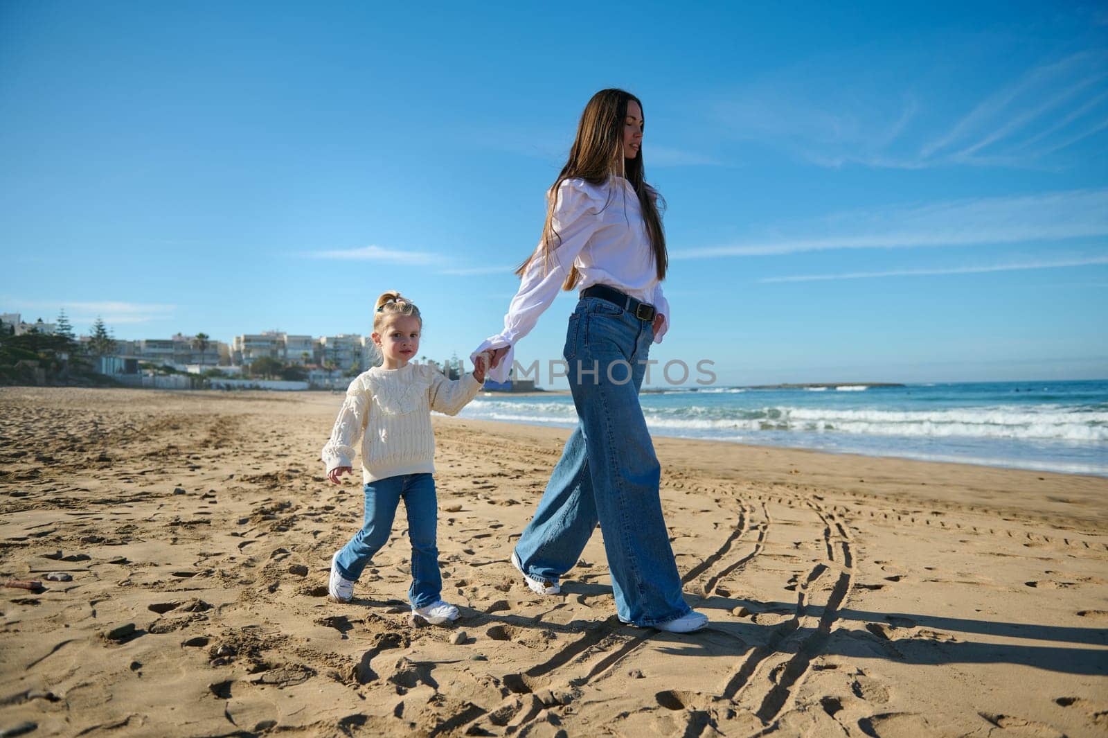 Full length portrait of a young mother and daughter, dressed in white shirts and blue jeans, holding hands and walking along the tropical beach, enjoying a happy weekend together outdoor