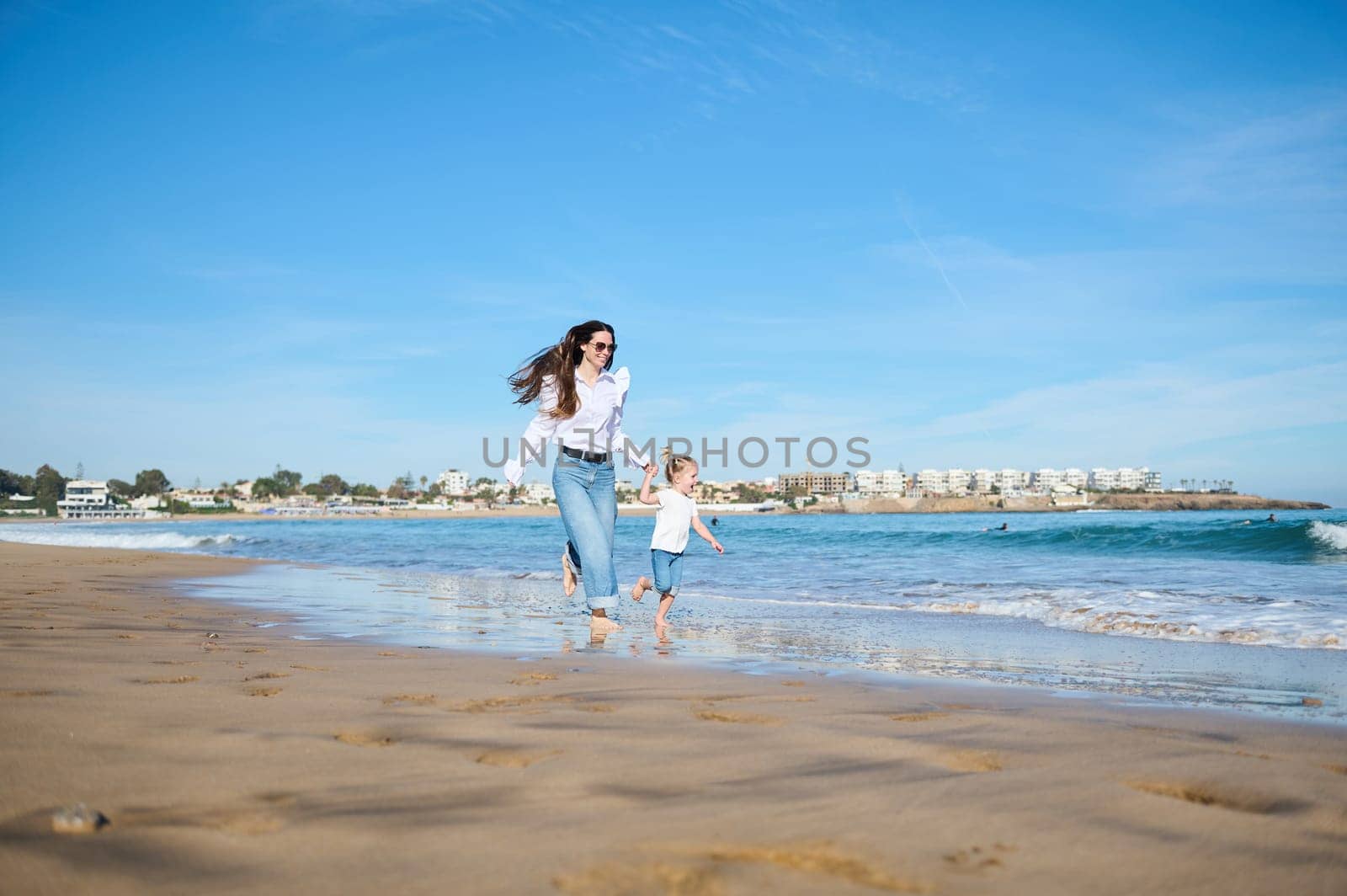 Mother and daughter girl running barefoot together on sandy beach in sea water in summer in warm ocean waves. by artgf