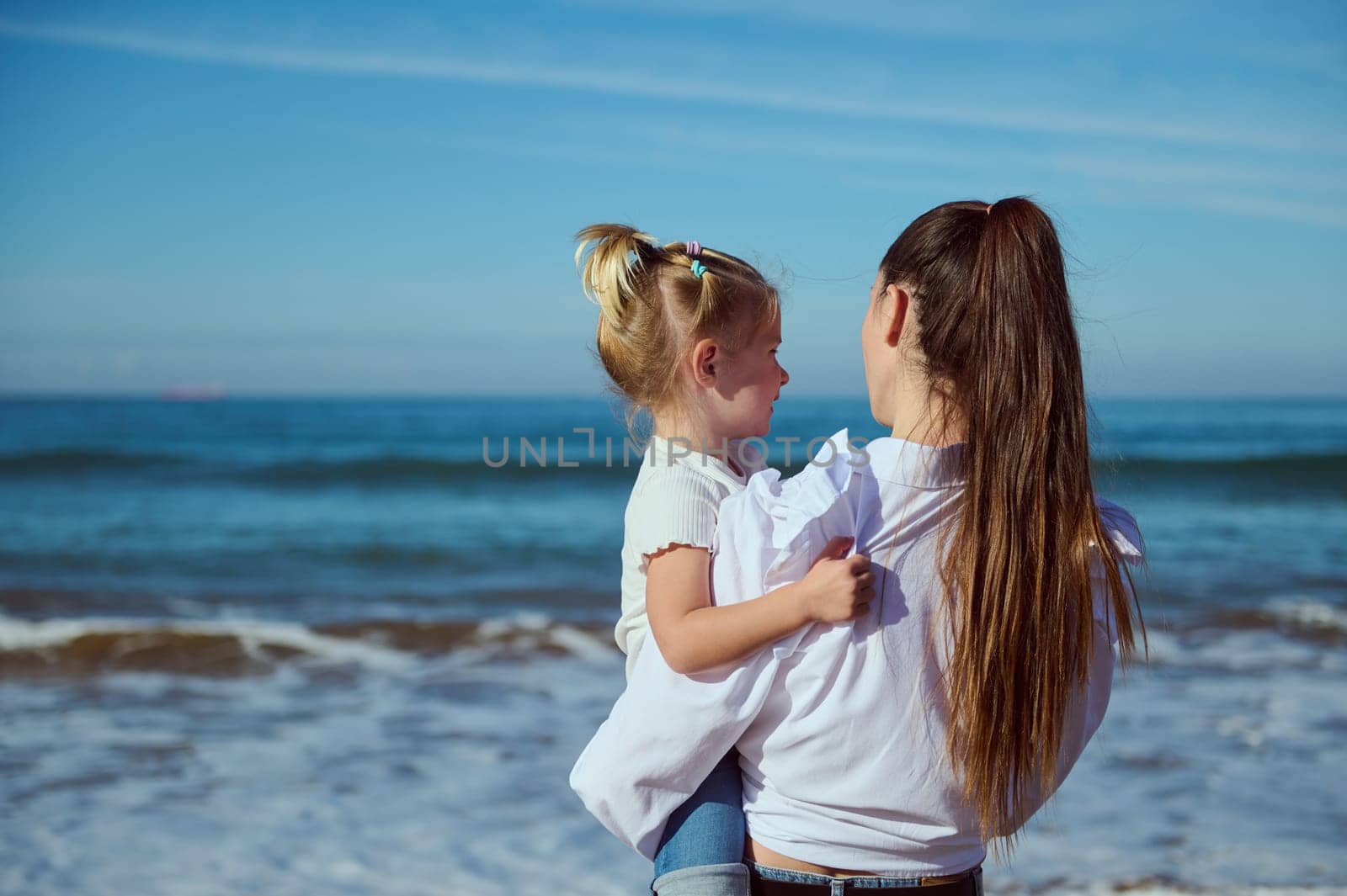 Rear view of a Caucasian mother holding her daughter on arms, standing together on the beach and admiring beautiful waves splashing while pounding on the sandy shore. People and nature. Family pastime