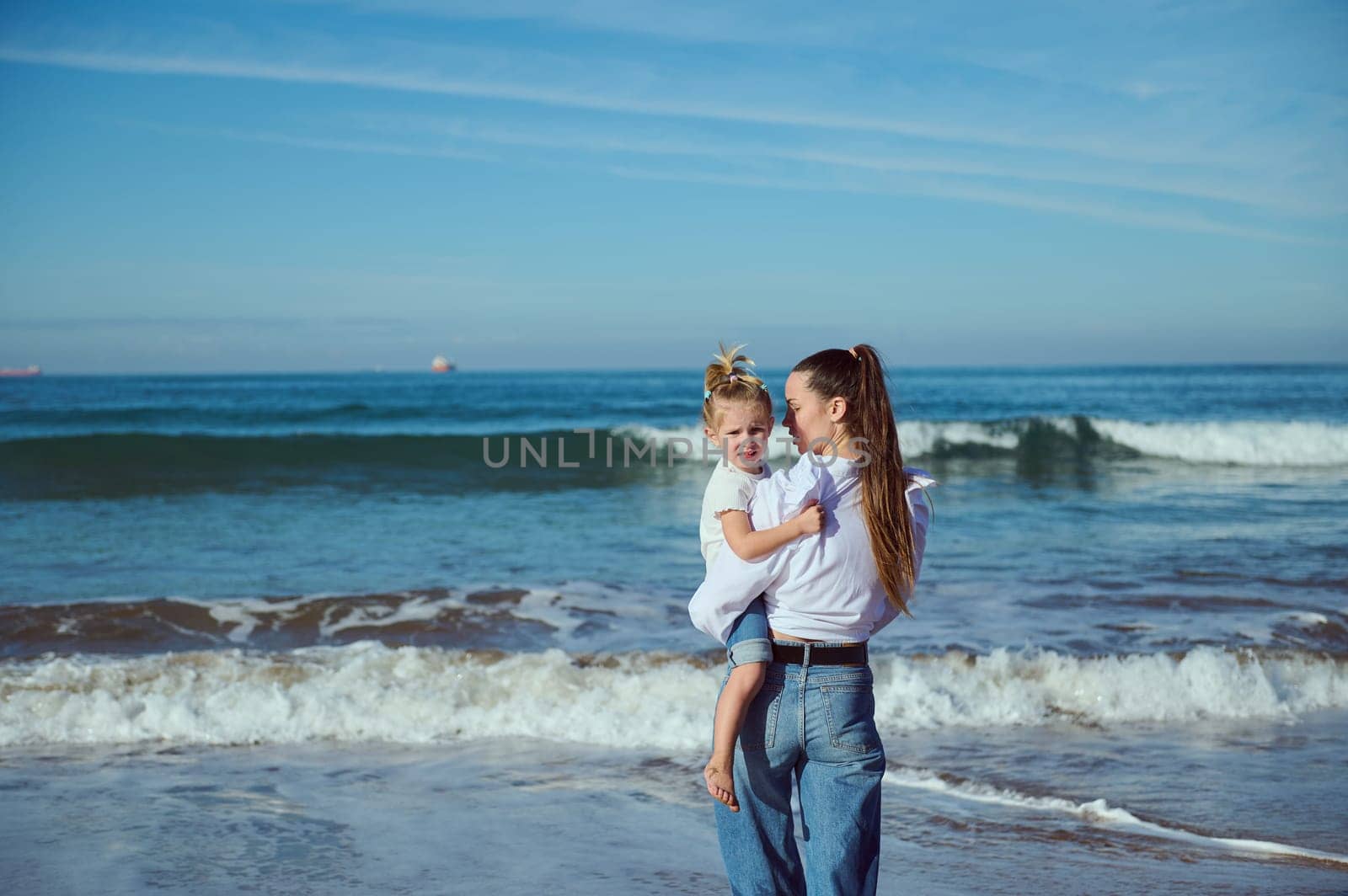 A beautiful young mother in white casual shirt and blue jeans, carries her baby in her arms, an adorable little girl, walking together barefoot on the beach. Mom and daughter. People and nature