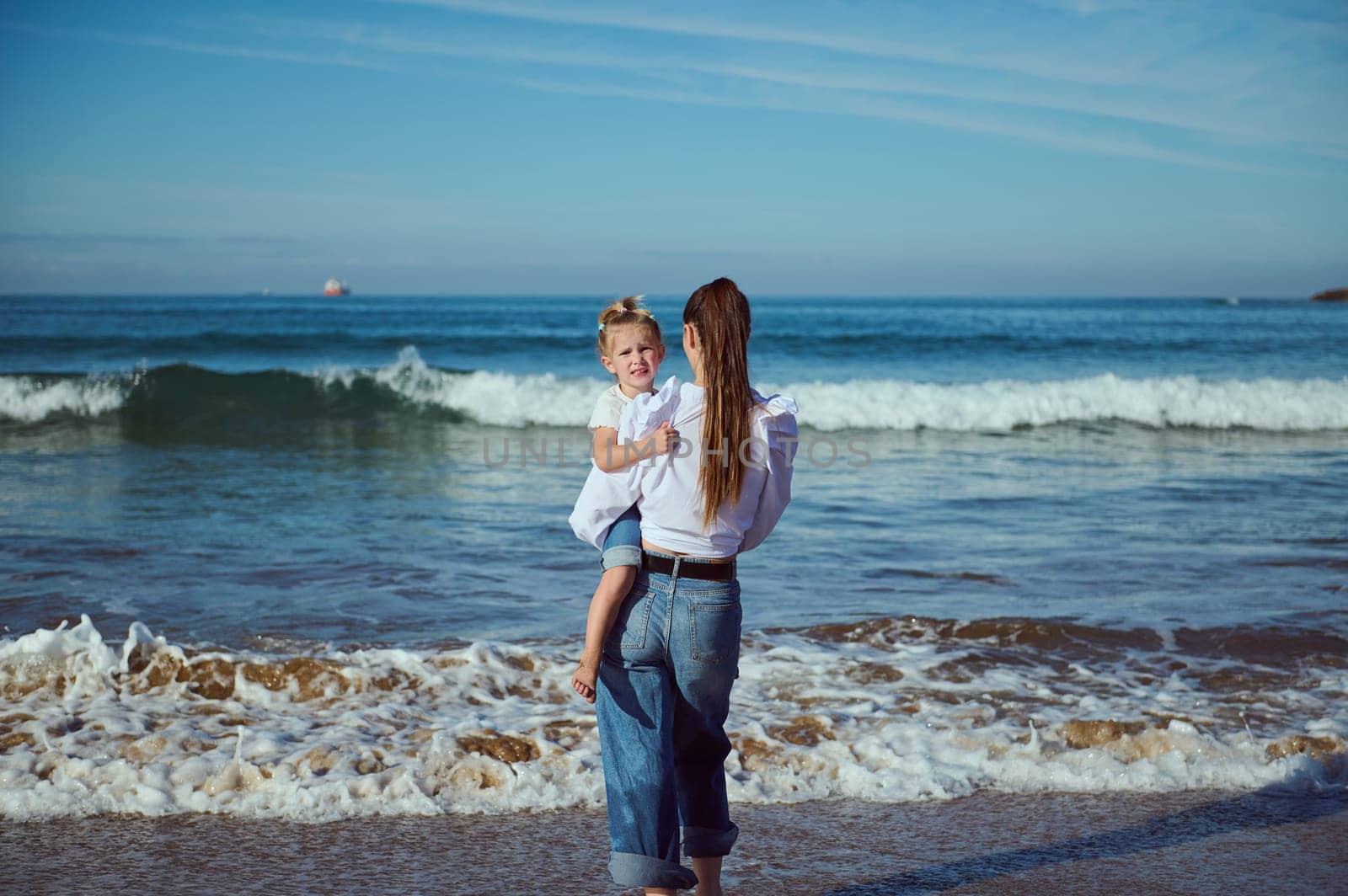 View from the back of a young mother carrying her daughter on the beach. Loving mom walking barefoot with her cute child on the sandy beach