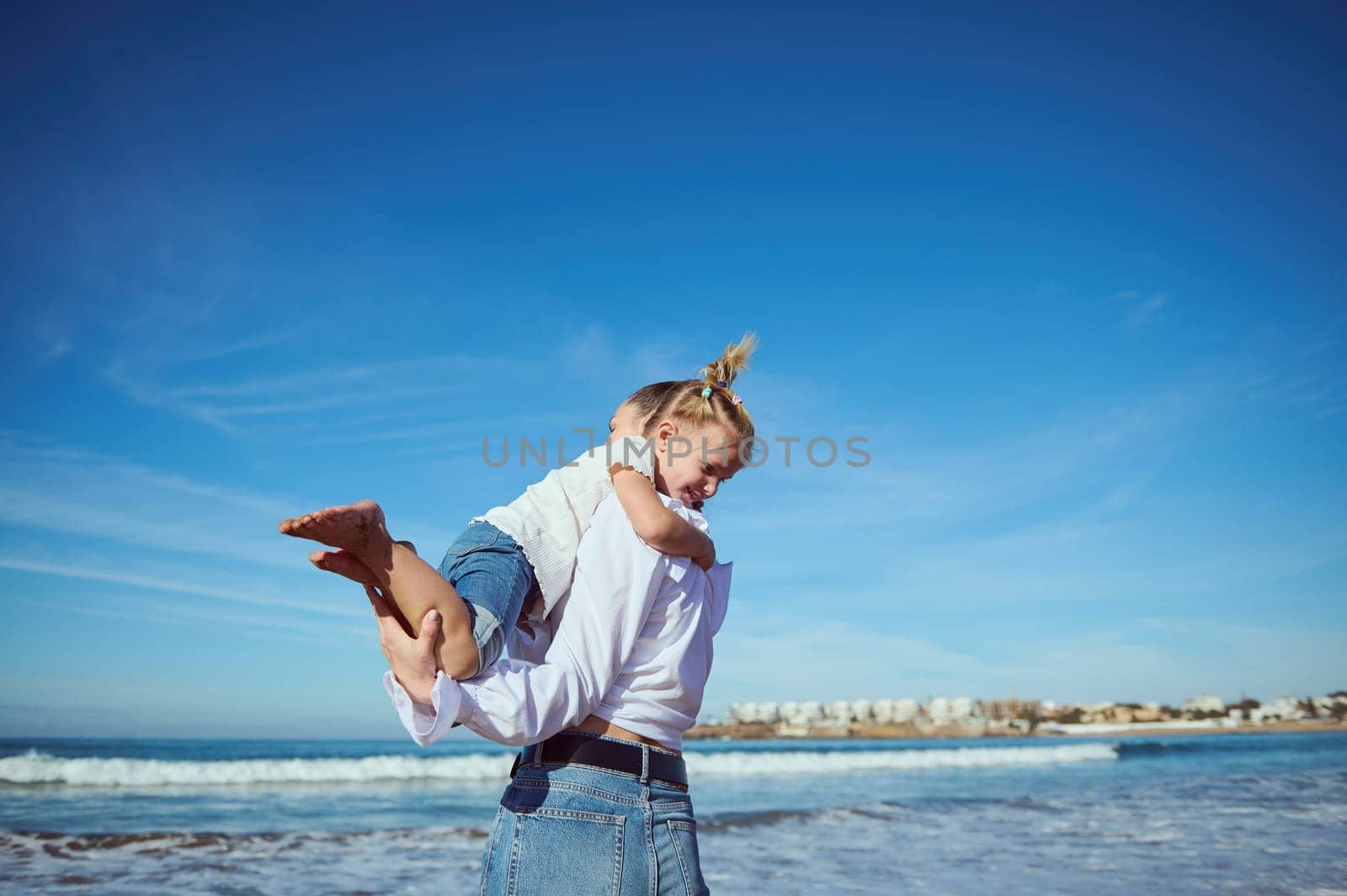 Authentic portrait of Caucasian pretty happy young woman, mother playing with her cute baby girl on the beach, turning around, laughing, walking leaving footstep on the wet sand. Family relationships