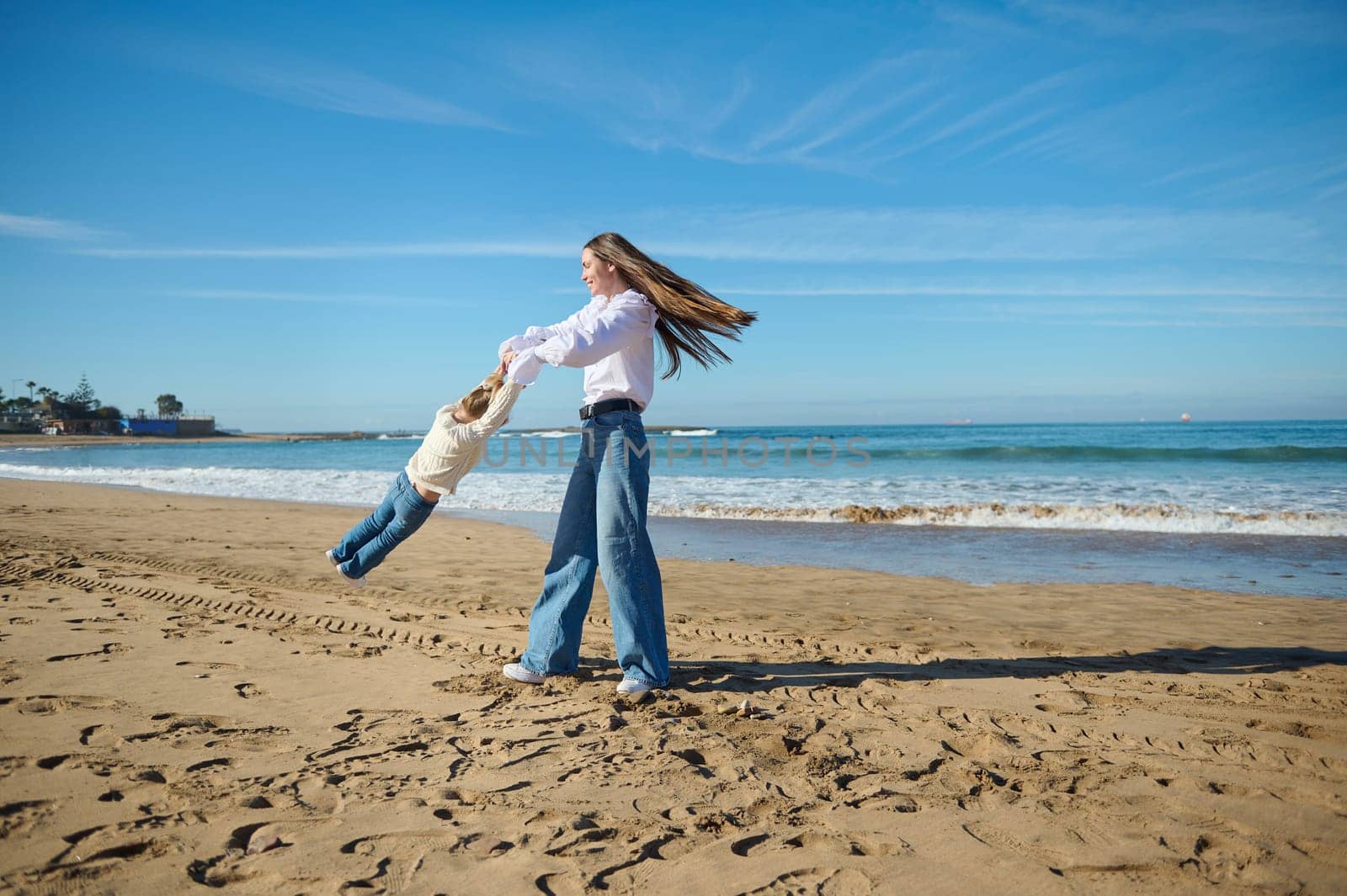 Full length view of a happy cheerful mother spinning daughter at beach, enjoying wonderful time together on the nature, standing on the sand against blue clear sky and Atlantic ocean background