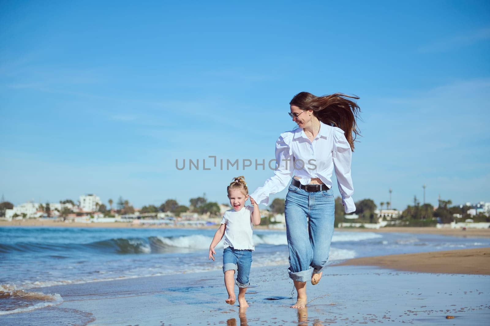 Caucasian happy young mother and daughter smiling while running on the beach, leaving footsteps on the wet sand by artgf