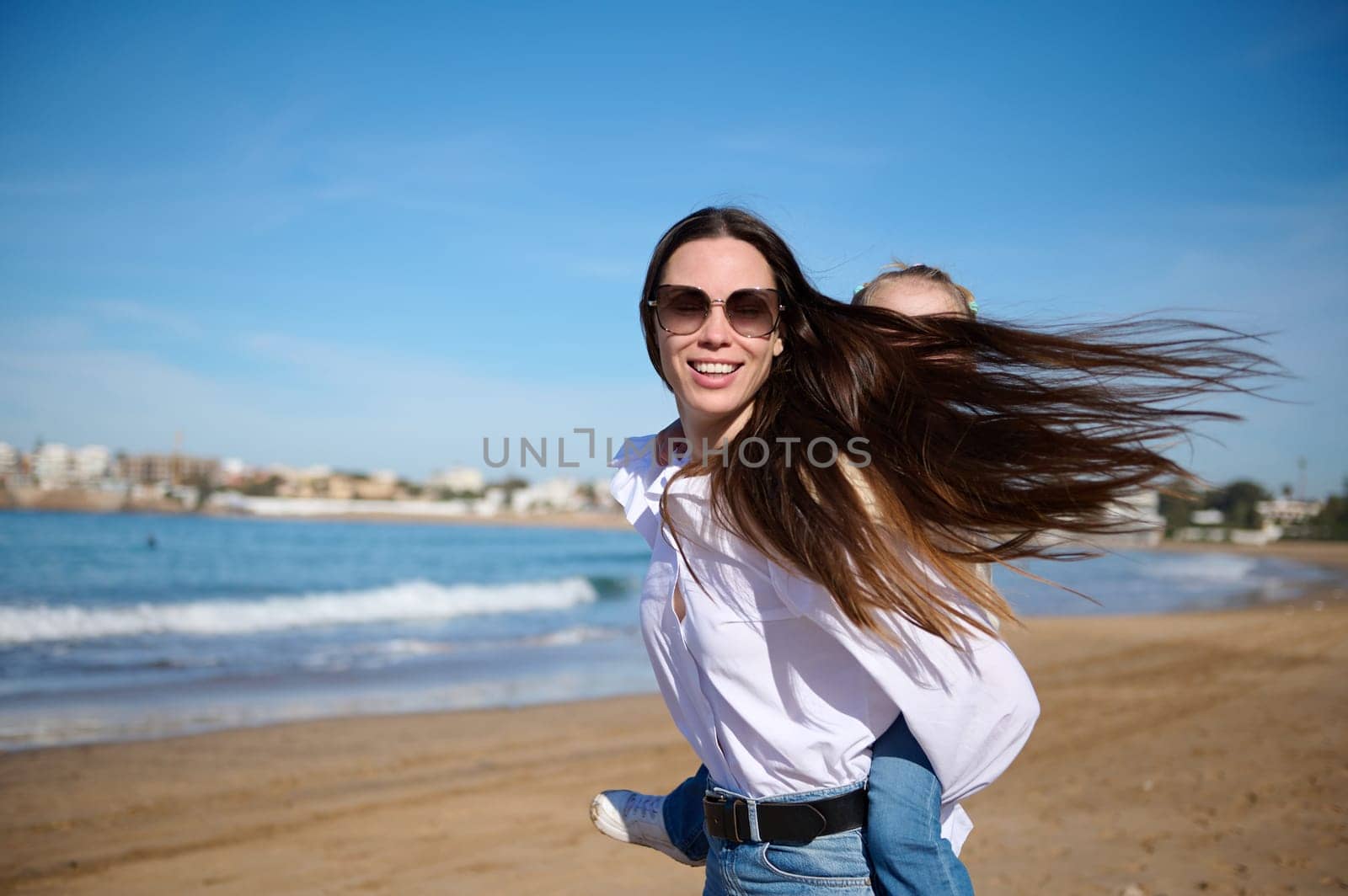 Caucasian young happy mother smiling looking at camera while giving piggyback ride to her lovely daughter, spinning her around herself and enjoying happy family time together on the Atlantic beach.