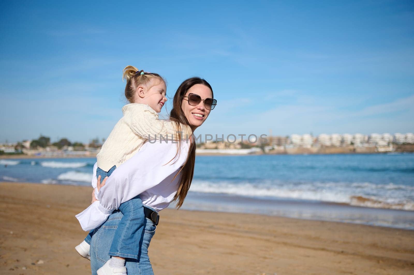 Happy mother and daughter playing on beach, carrying kid hugging, piggy back fun games, looking smiling outdoors. Family bonding together on summer holiday, travel activities recreation lifestyle.