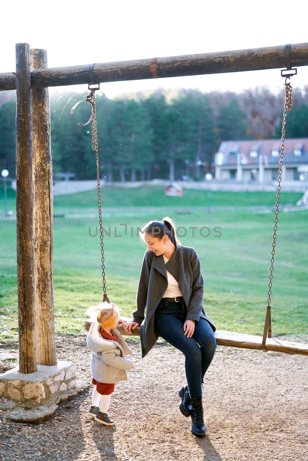 Little girl stands next to her mom sitting on a wooden chain swing in the park. High quality photo