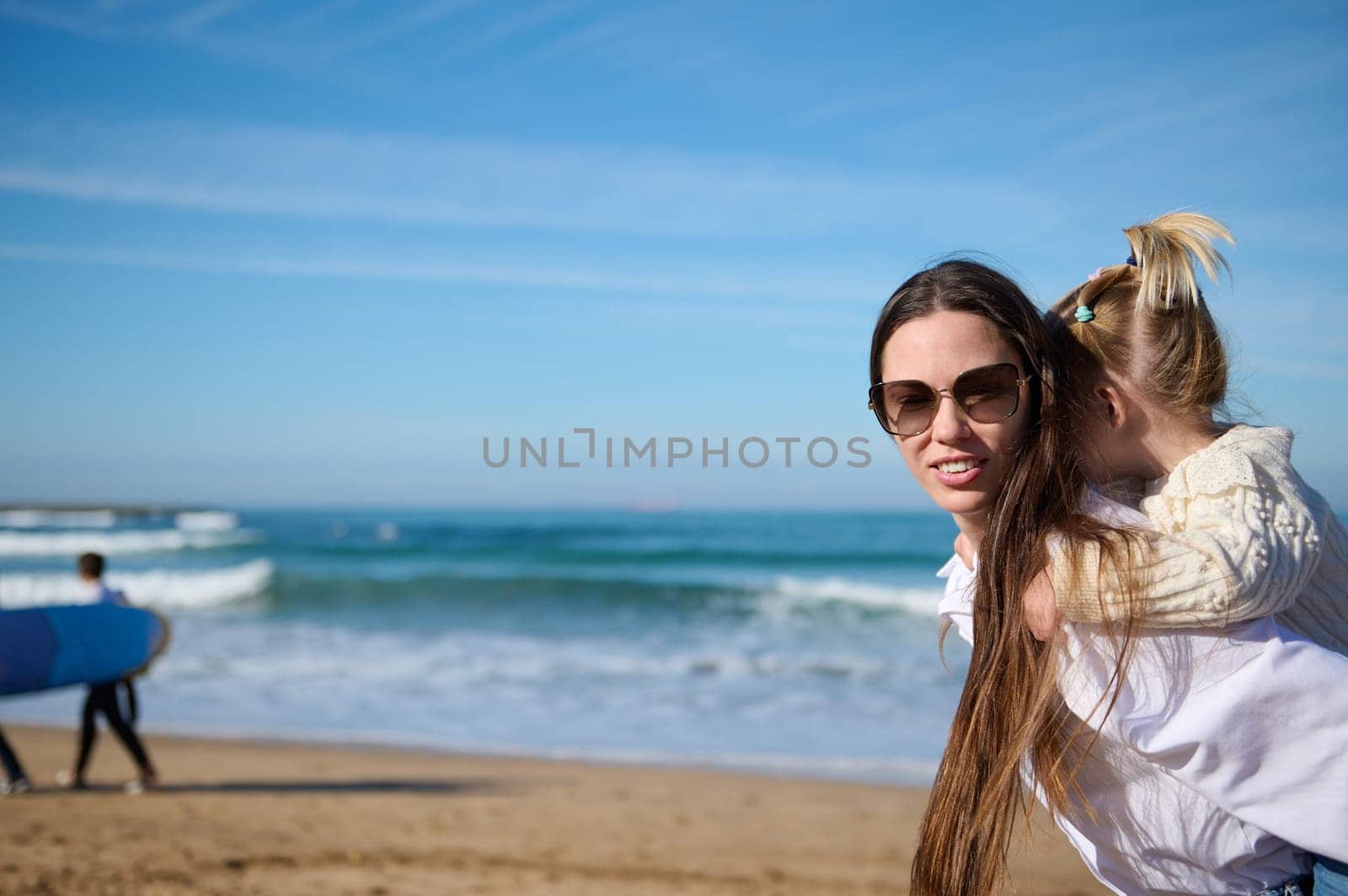 Happy young woman, mother and lovely daughter having fun together, walking on the beach against unrecognizable people practicing surf on the Atlantic beach. People. lifestyle. Leisure activity