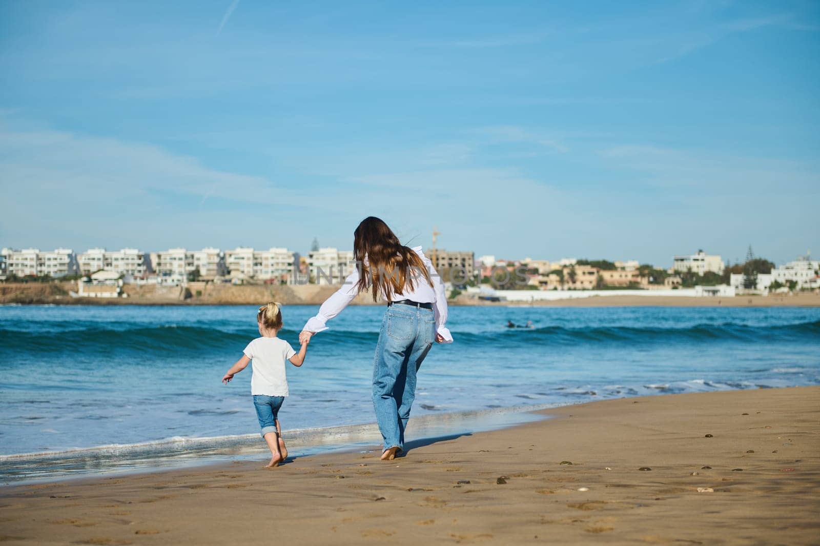 Caucasian blonde little child girl holding her mother's hand, while walking in the warm water along the sea, enjoying the waves washing her feet. People and nature. Active healthy lifestyle