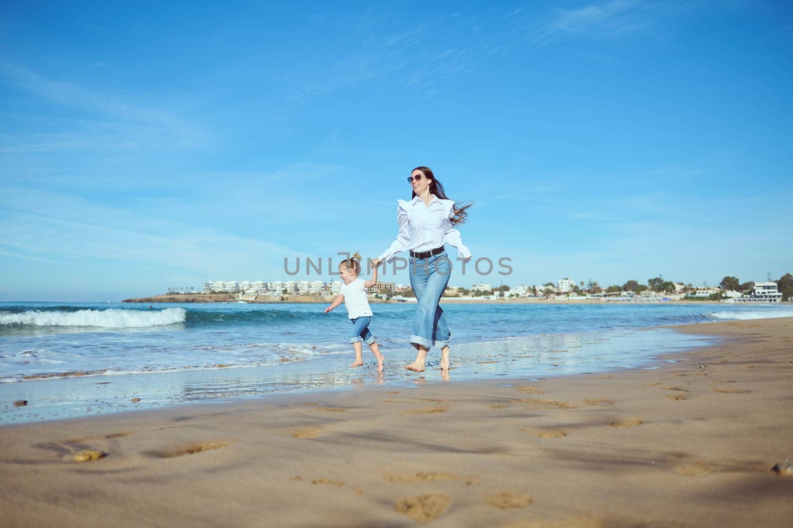 Mother and daughter holding hands, running barefoot on the wet sand, leaving footsteps, enjoying happy moments together by artgf
