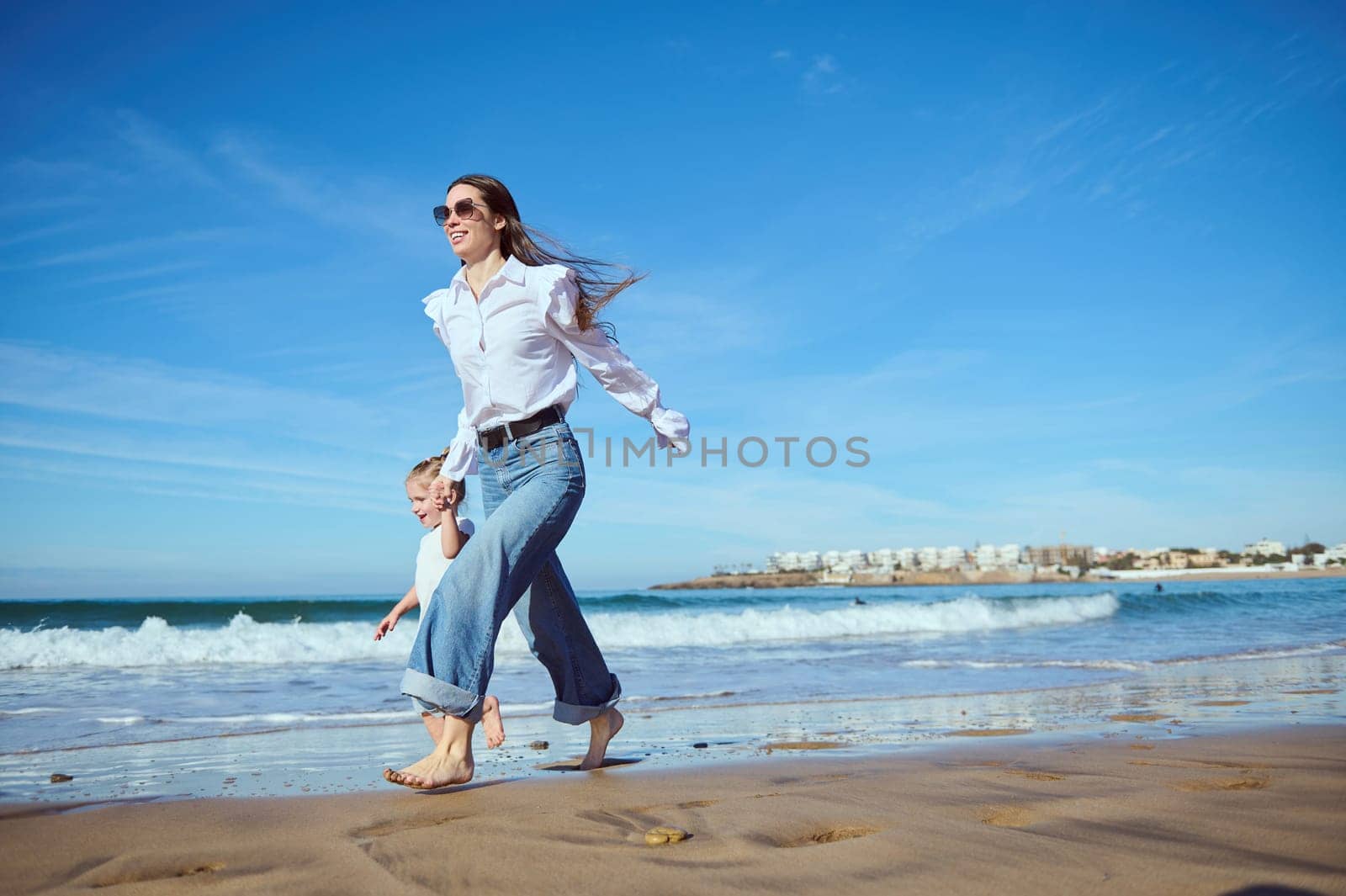 Beautiful woman, happy loving mom holds the hand of her cute little child girl while running walking in the warm water along the sea, enjoying the waves washing her feet. People and nature. Lifestyle