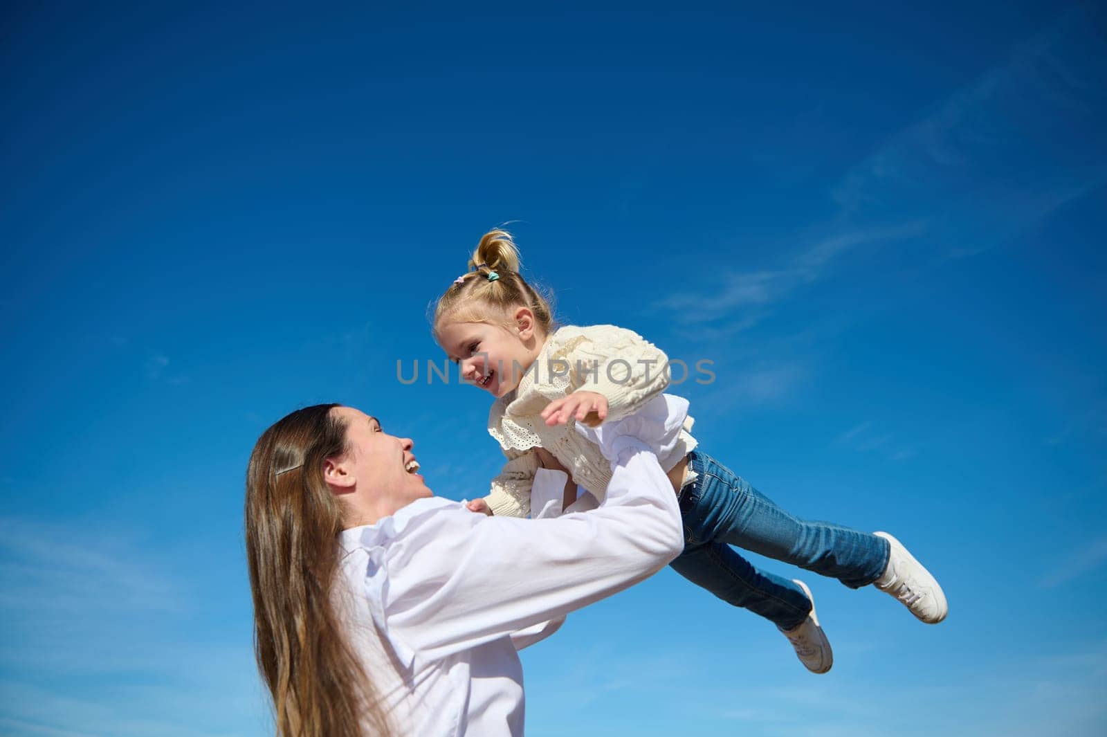 Bottom view of a happy harmonious family of a young loving caring mother throwing up her little baby, a cute child girl, laughing and playing in the summer on the nature against blue sky background