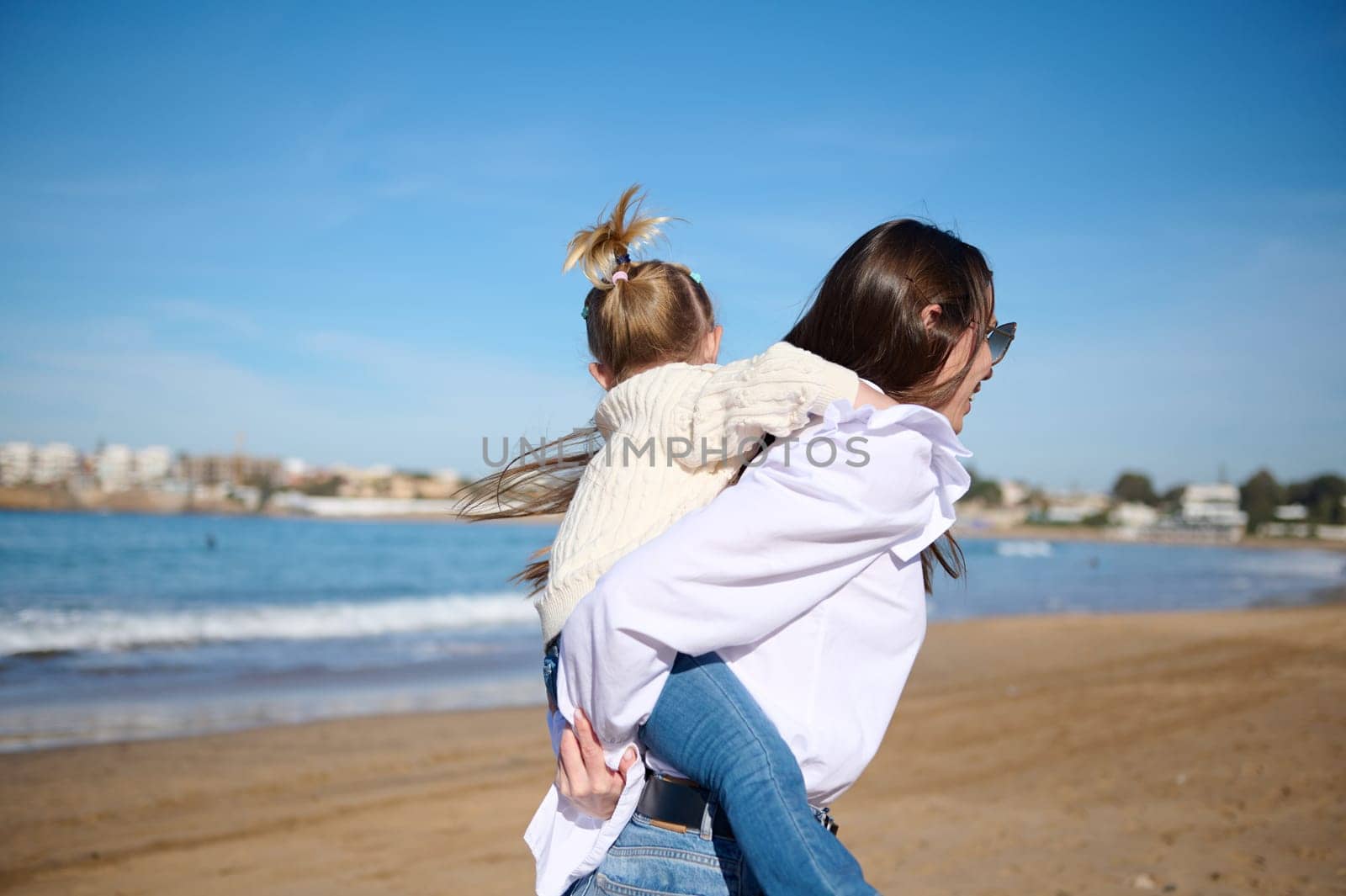 Rear view of a happy mother giving piggyback ride to her lovely daughter, spinning her around herself and enjoying happy family time together on the Atlantic beach.