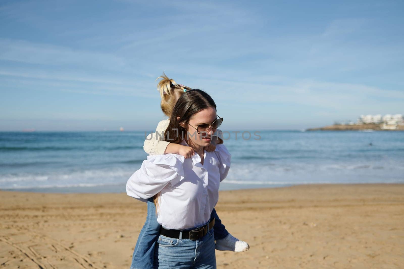 Caucasian young woman, mother in white l shirt, blue jeans and sunglasses, carrying her little daughter on her back, walking on the sandy beach along the beautiful waves breaking on the Atlantic shore