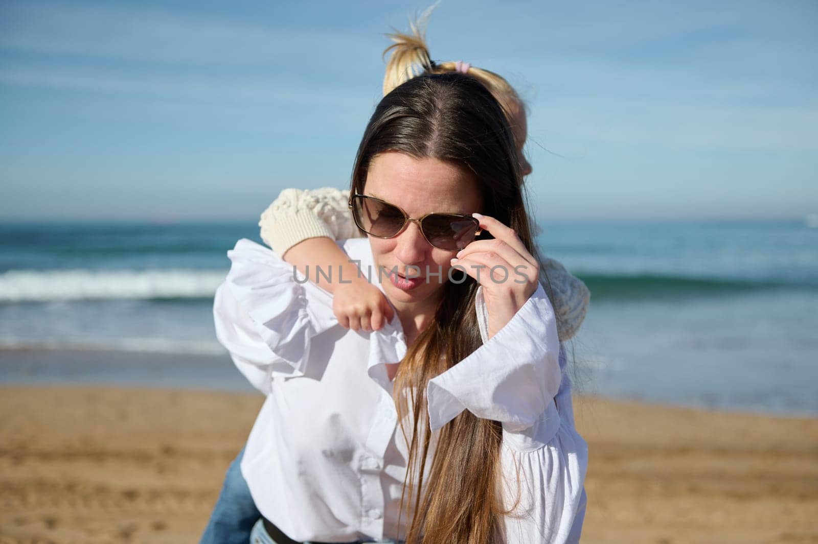 Portrait of a brunette woman, young loving caring mother in eyeglasses, carrying her little daughter on her back while spending happy time together on the beach. Family. Motherhood. Carefree childhood