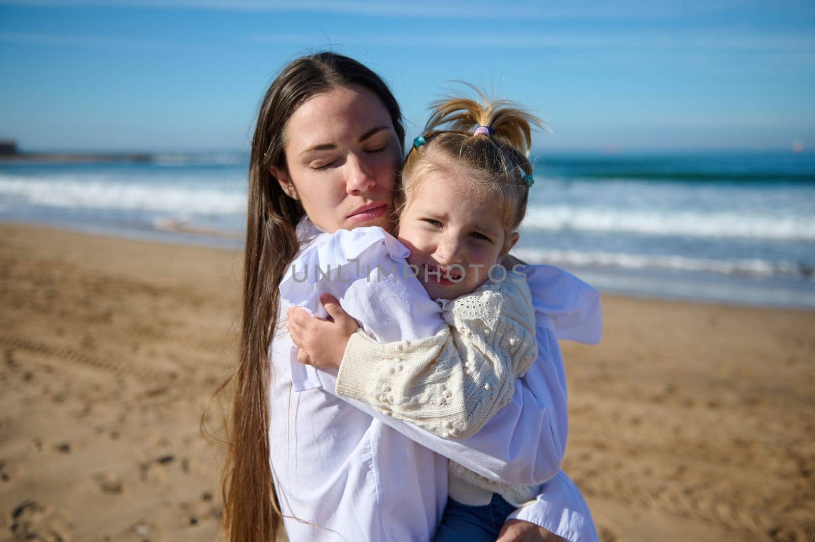Caucasian delightful young mother gently hugging her little kid girl, standing with her eyes closed on the sandy beach, against the background of beautiful waves pounding on the Atlantic Sea shore