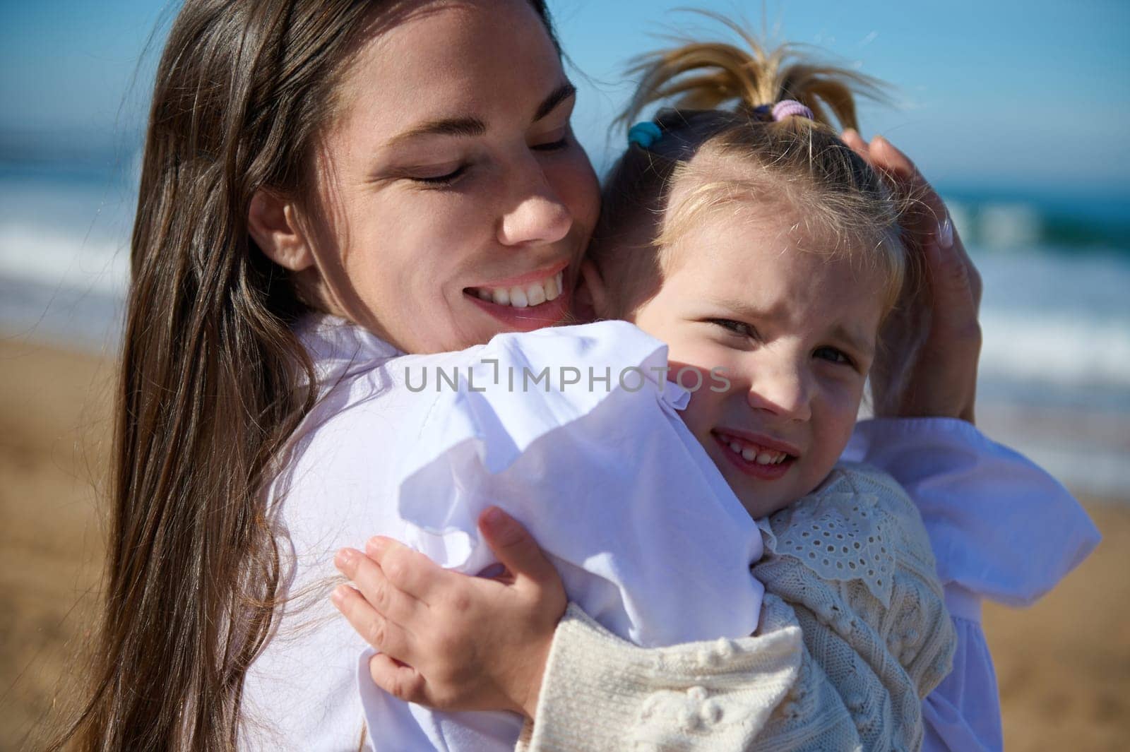 Close up portrait of authentic happy woman, young mother smiling and hugging her adorable little child girl, standing on the sandy beach together. People. Happy carefree childhood. Maternity concept