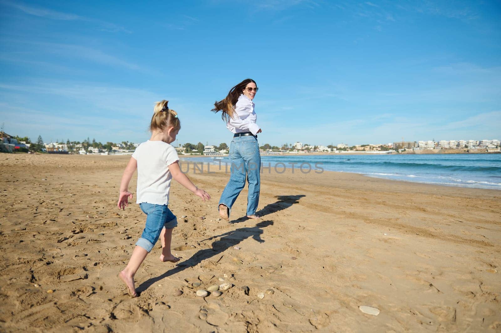 Rear view of a happy young woman, loving mother and her little daughter, playing together on the beach, running barefoot and leaving footsteps on the wet sand, enjoying a happy moments together