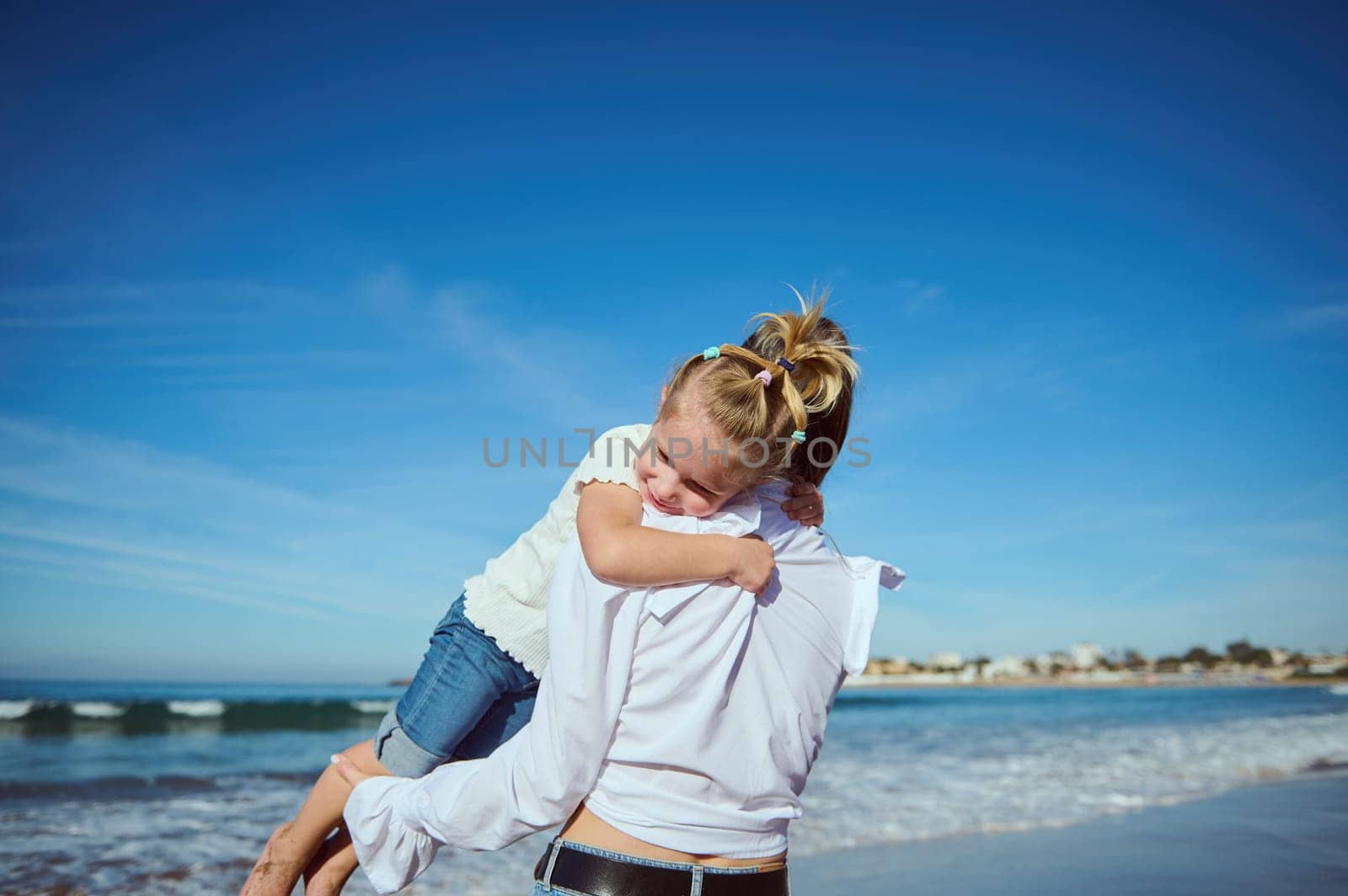 Adorable little kid girl daughter gentle hugging her mother who holds her in her arms while walking together on the sandy beach. Happy carefree childhood. Maternity leave. Family relationships concept