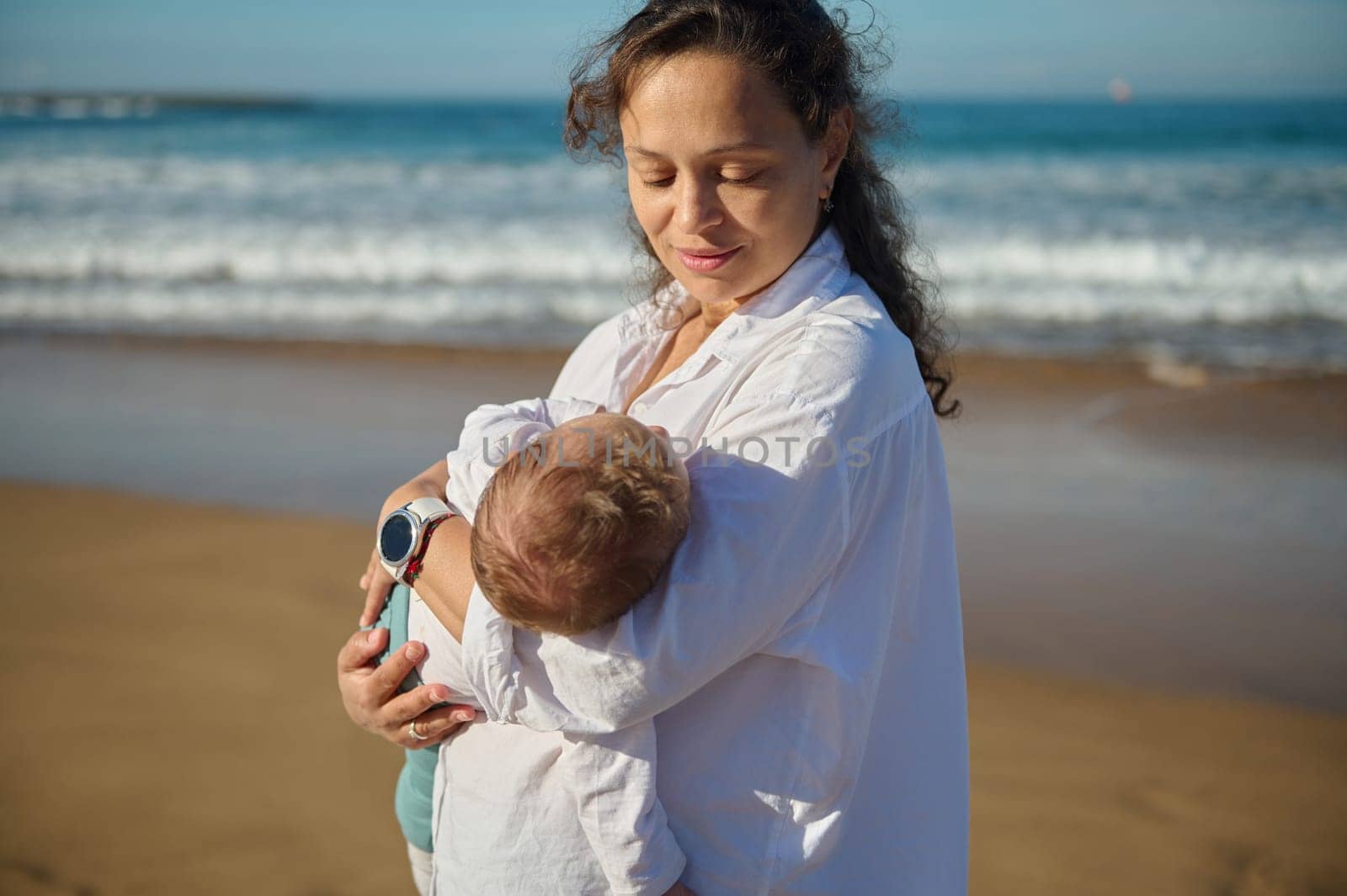 Beautiful curly brunette, loving caring mother holding her baby boy, rocking and kissing him while he's falling asleep in her arms, walking along the beach together on a beautiful warm sunny day