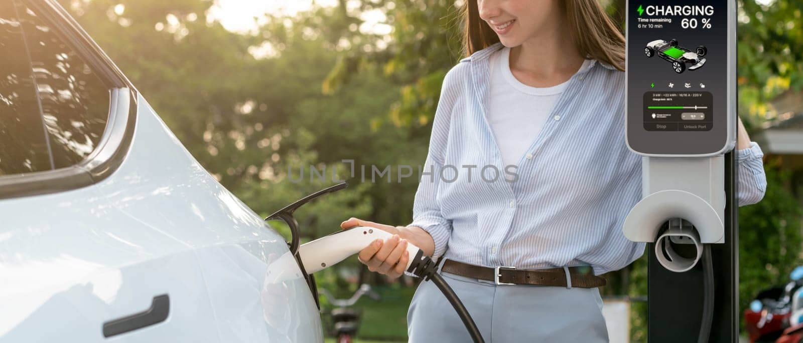 Young woman recharging battery for electric car during road trip travel EV car in natural forest or national park with sunrise lighting. Eco friendly travel during vacation and holiday. Panorama Exalt